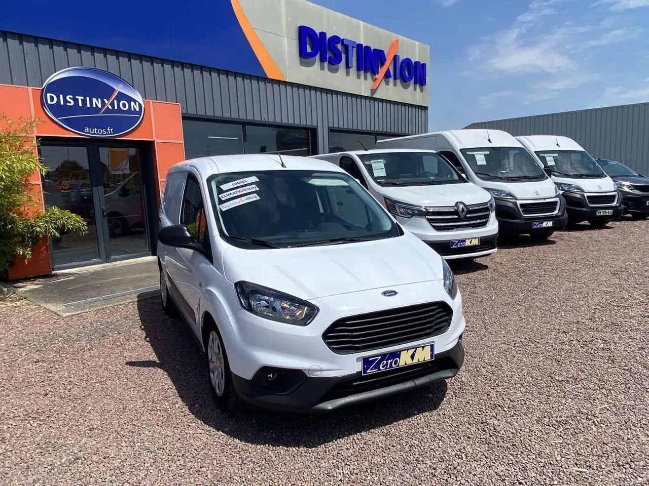 Vue 3/4 avant d'un Ford Tourneo Courier blanc stationné devant un concessionnaire, avec calandre noire et enjoliveurs visibles.