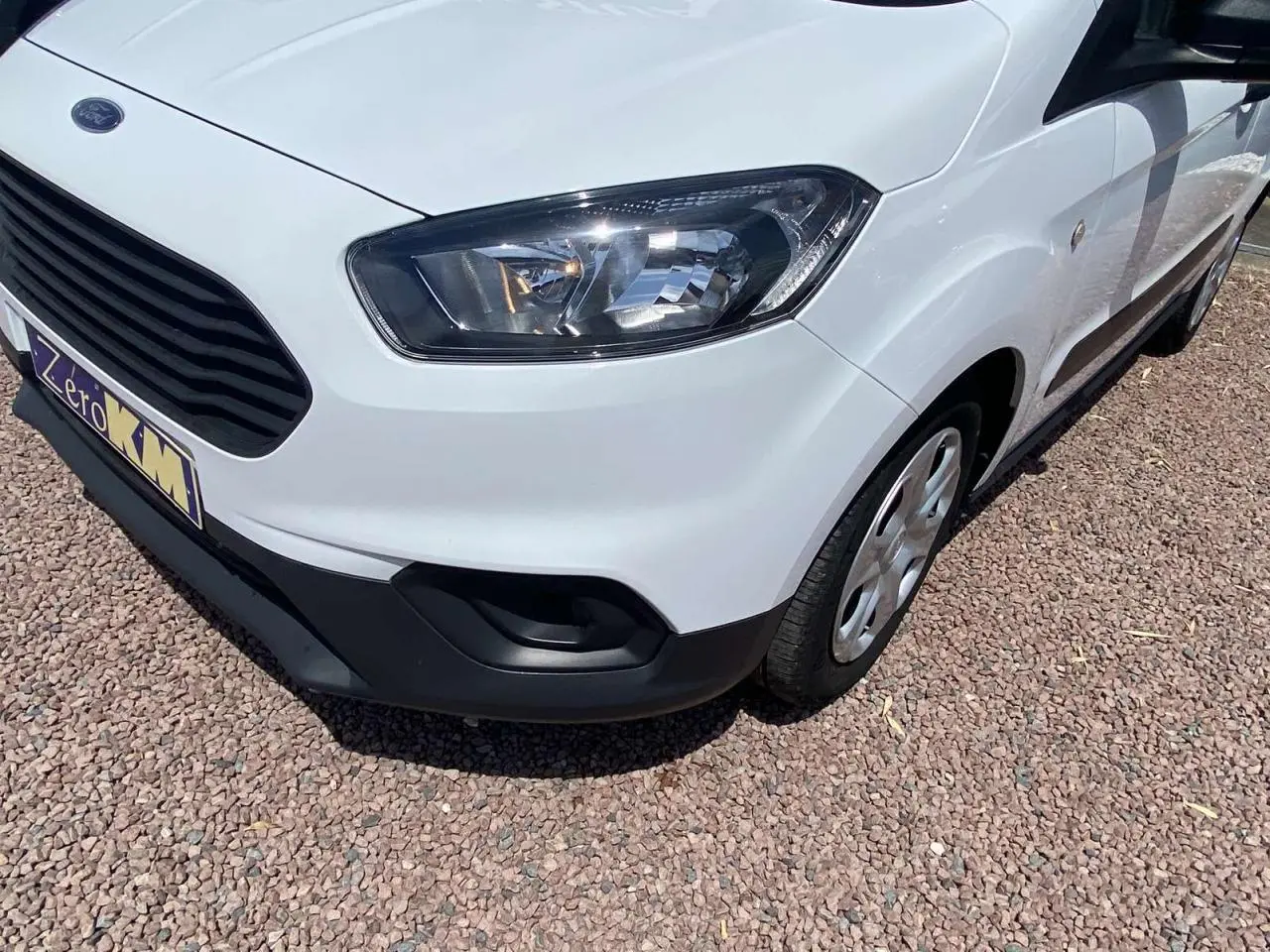 Vue 3/4 avant droit d'un Ford Tourneo Courier blanc avec enjoliveurs cache moyeu sur sol gravillonné.