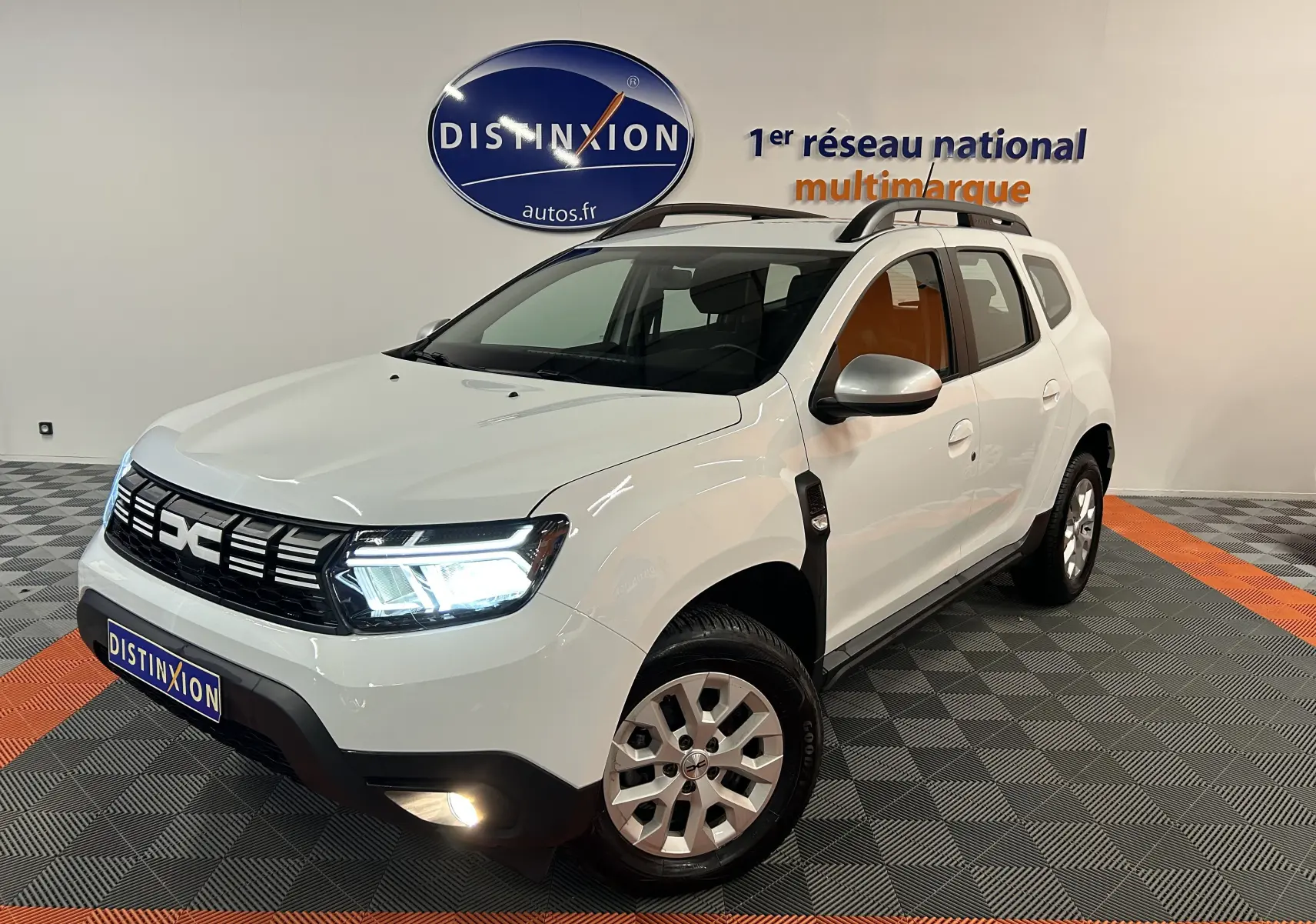 Vue 3/4 avant droite d'un Dacia Duster blanc 2023 avec feux allumés et barres de toit noires en intérieur showroom.