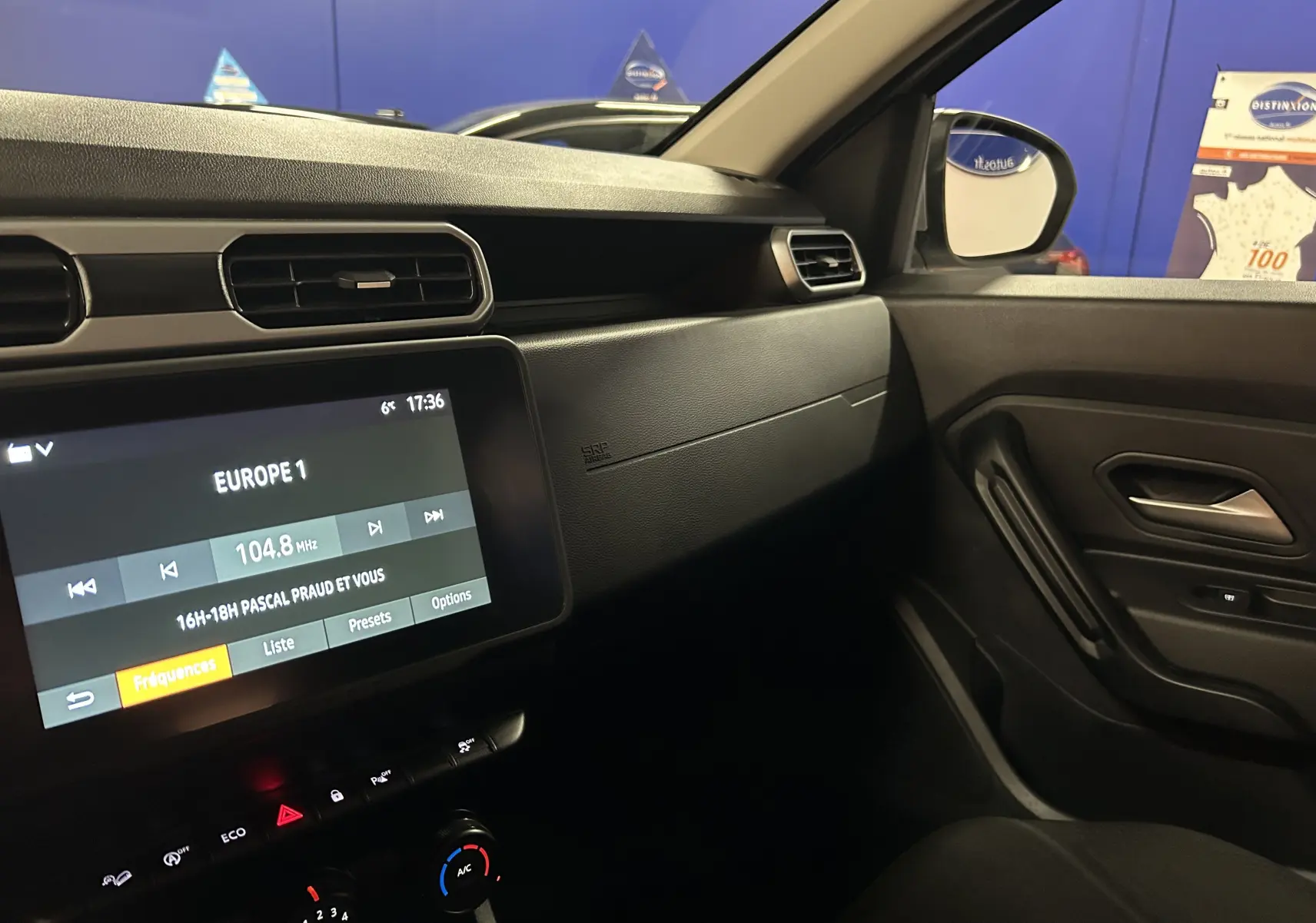 Vue intérieure côté passager du tableau de bord noir du Dacia Duster 2023 avec écran tactile et commandes climatisation.