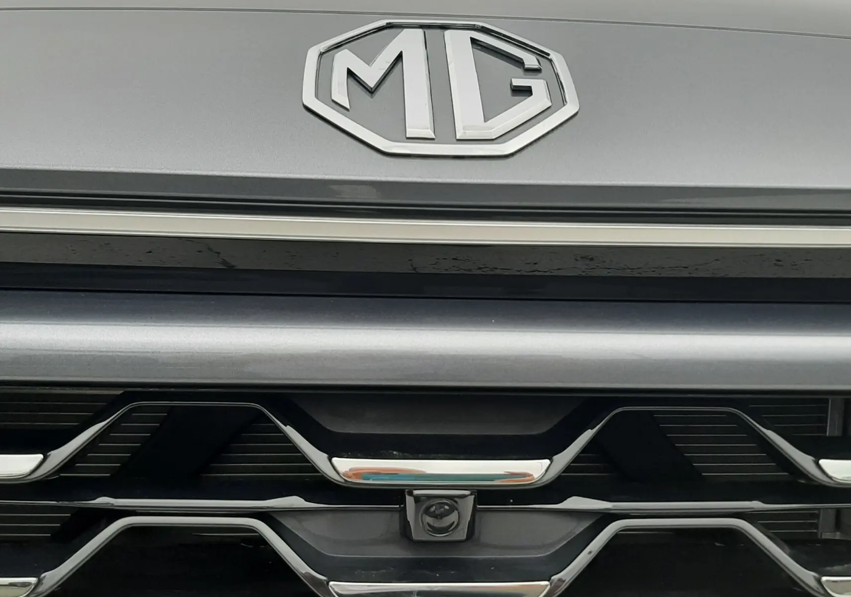Gros plan sur la calandre avant grise Camden Grey du MG ZS 1.5 Hybrid+ avec logo MG et caméra frontale intégrée.