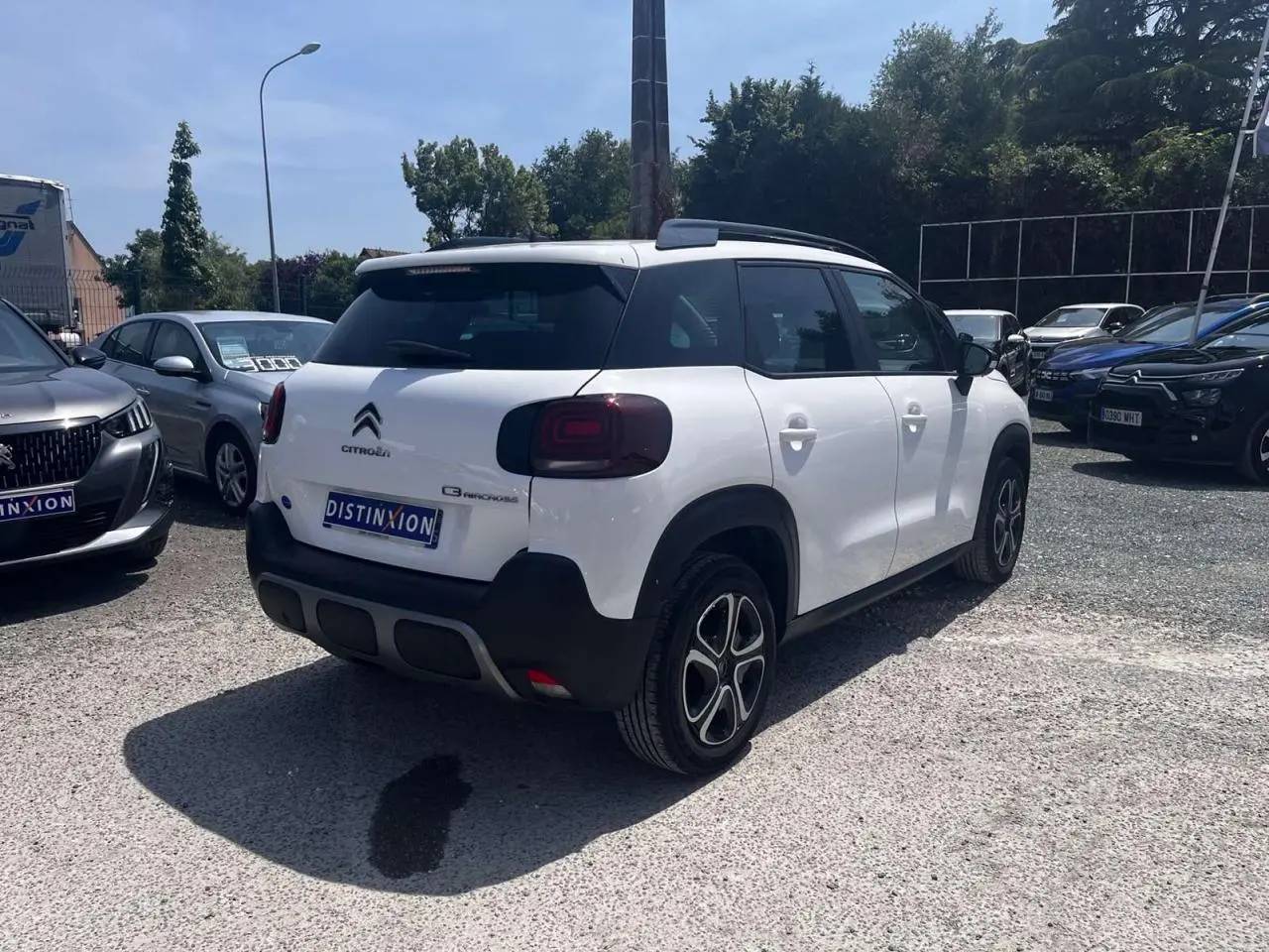 Vue 3/4 arrière droite d'un Citroën C3 Aircross blanc banquise avec barres de toit noir brillant sur parking.