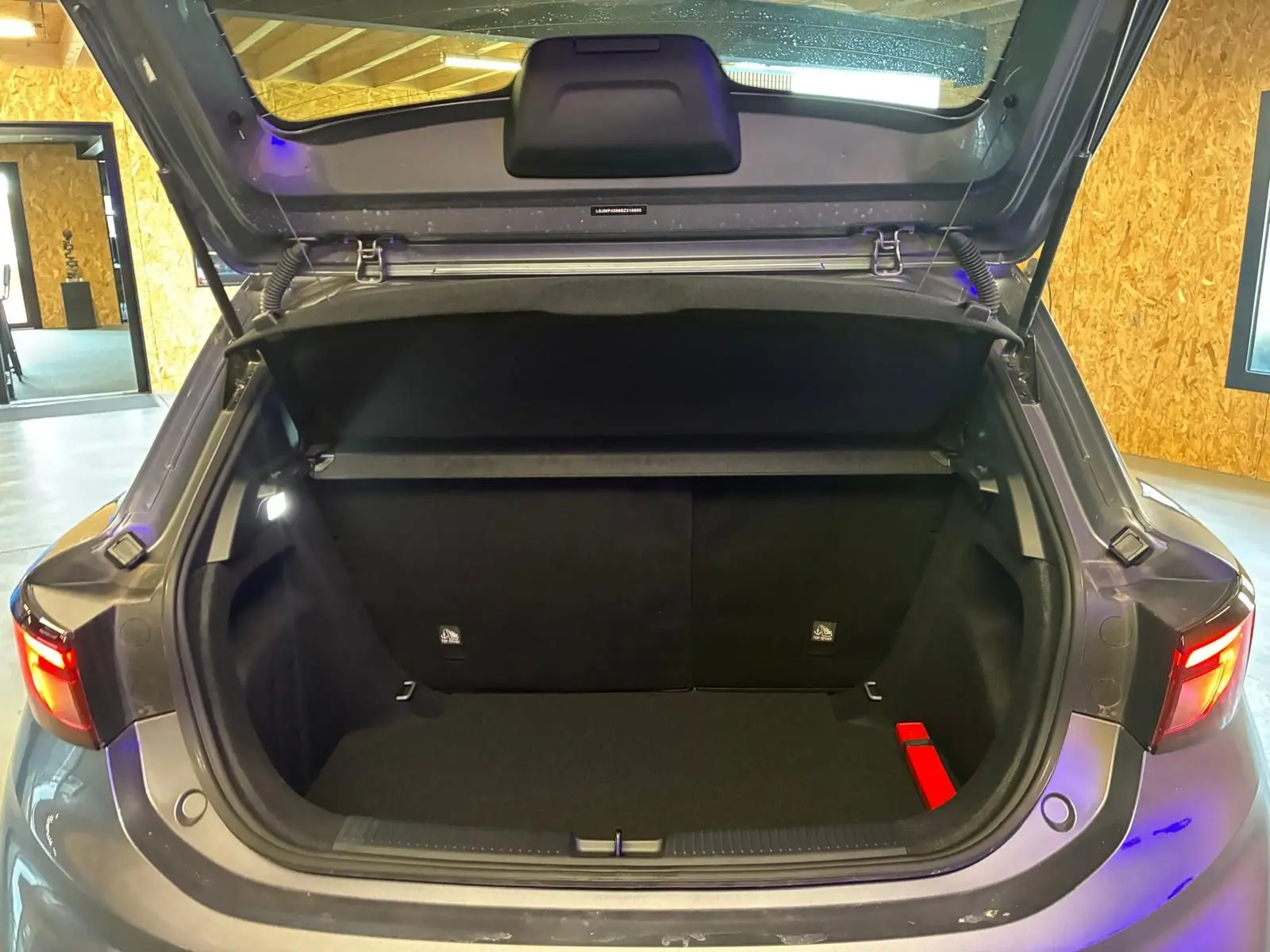 Coffre ouvert d'une MG3 1.5 HYBRID+ noire, vue arrière, avec feux arrière allumés dans un garage.