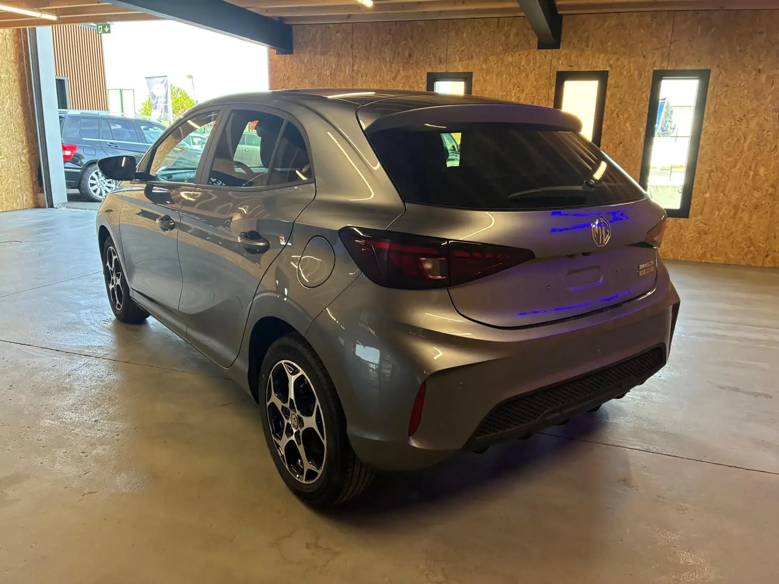 Vue 3/4 arrière droite d'une MG3 1.5 HYBRID+ 195 Luxury gris métallisé avec jantes noires et toit noir en intérieur showroom.