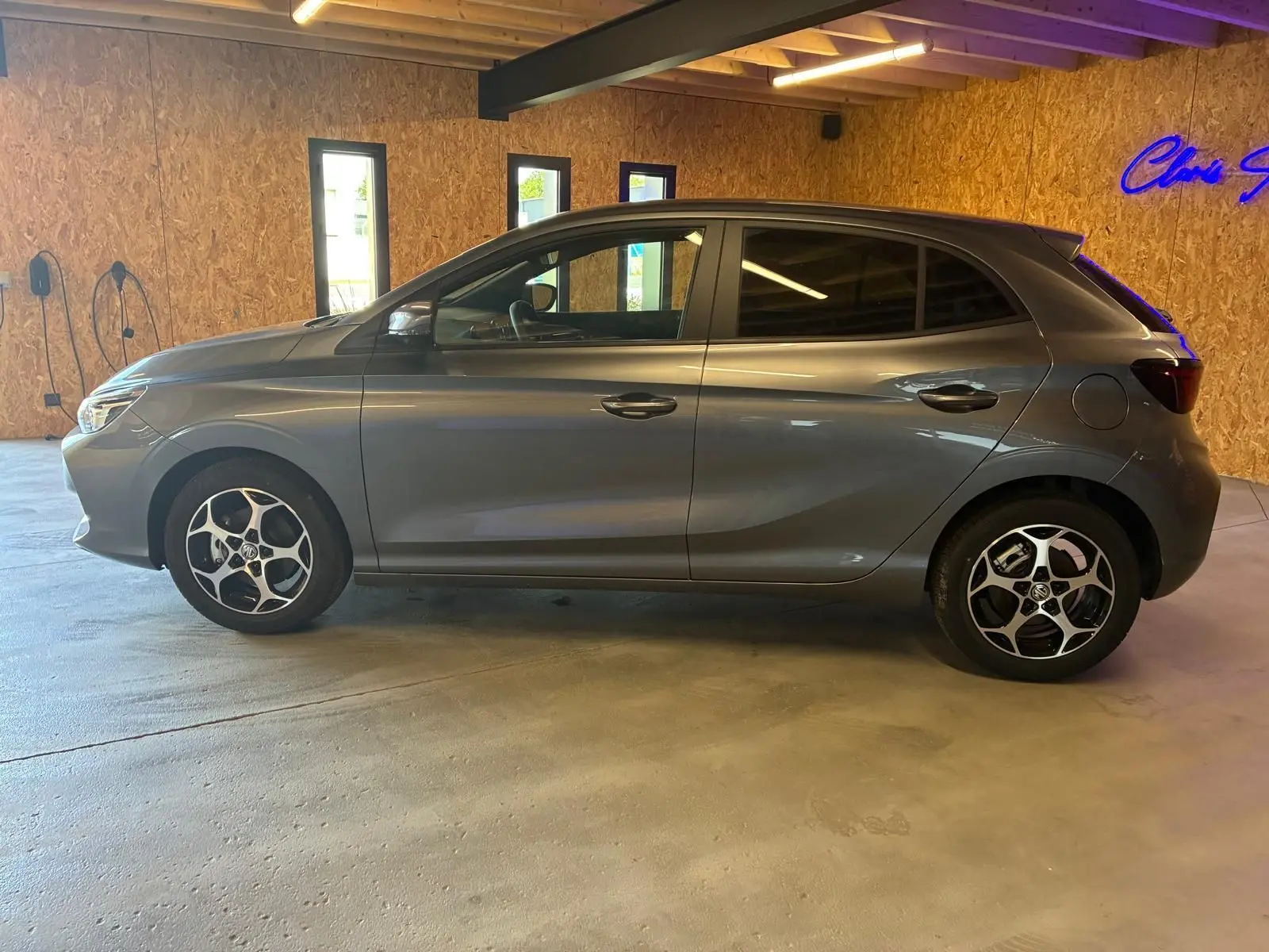Vue latérale droite du MG3 1.5 HYBRID+ 195 LUXURY gris métallisé avec jantes alliage et vitres teintées en intérieur showroom.