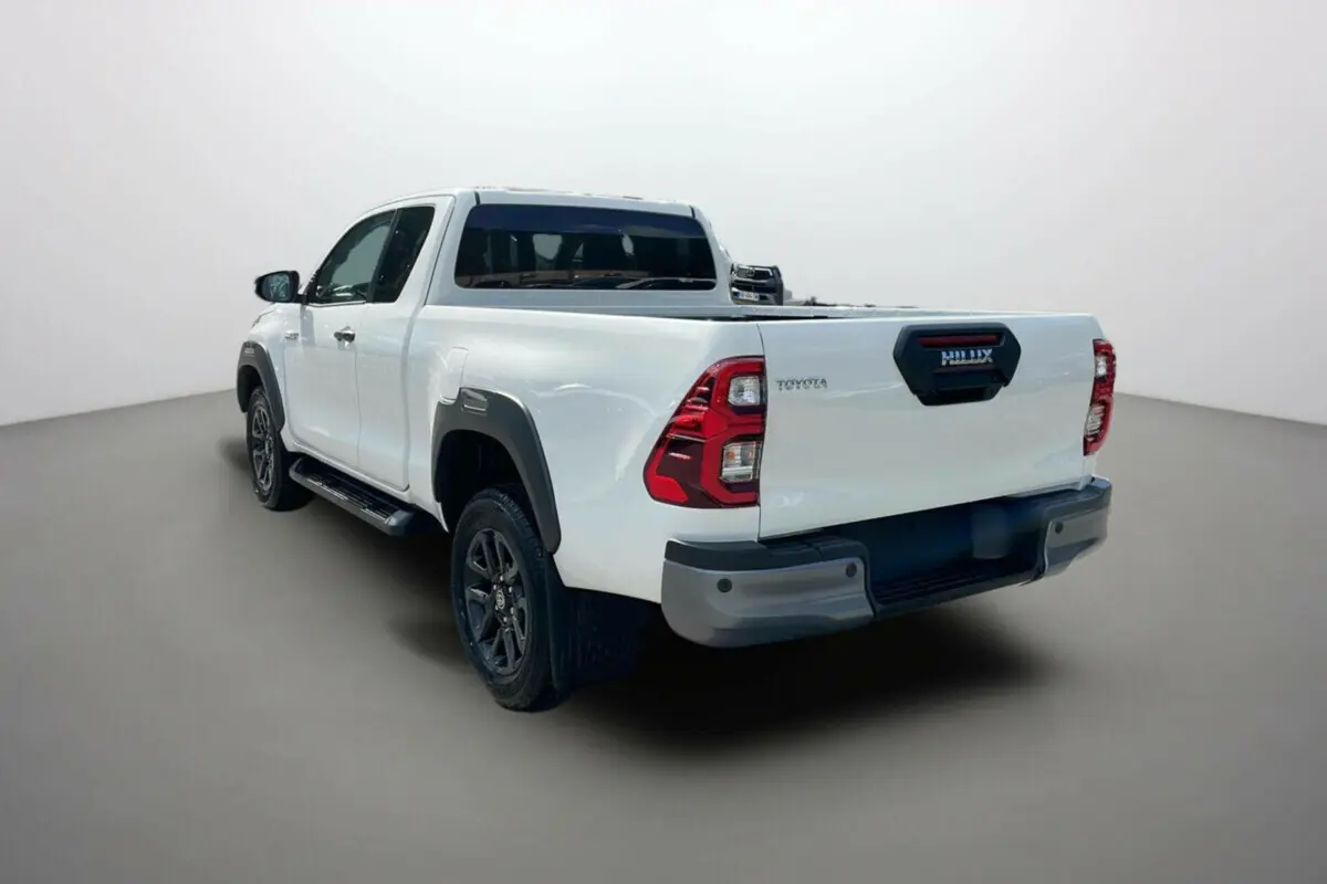 Toyota Hilux 4x4 blanc pur en 3/4 arrière droit, avec protections noires et jantes foncées visibles.