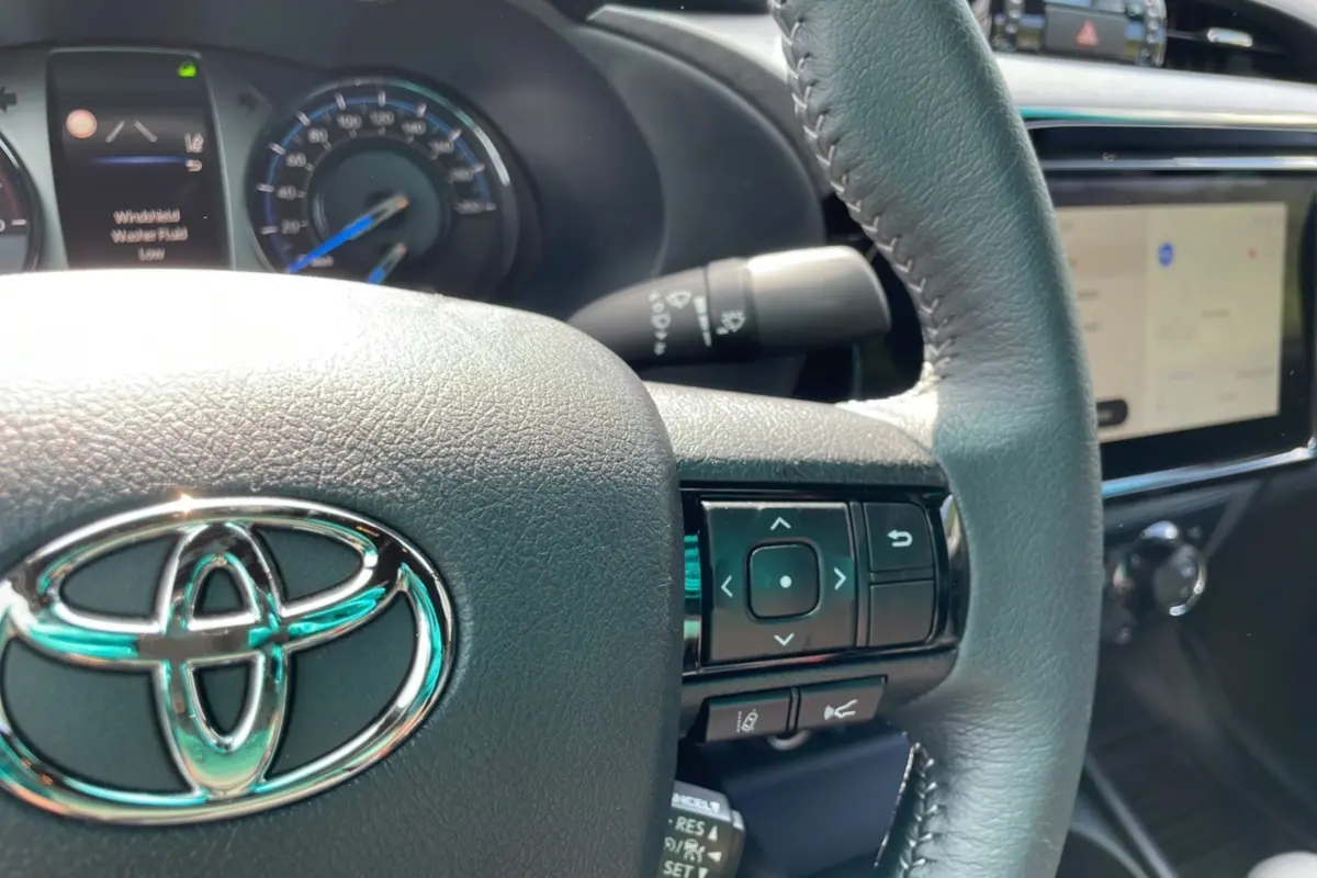 Vue rapprochée du volant cuir noir du Toyota Hilux 2025 blanc, avec commandes intégrées et tableau de bord numérique.