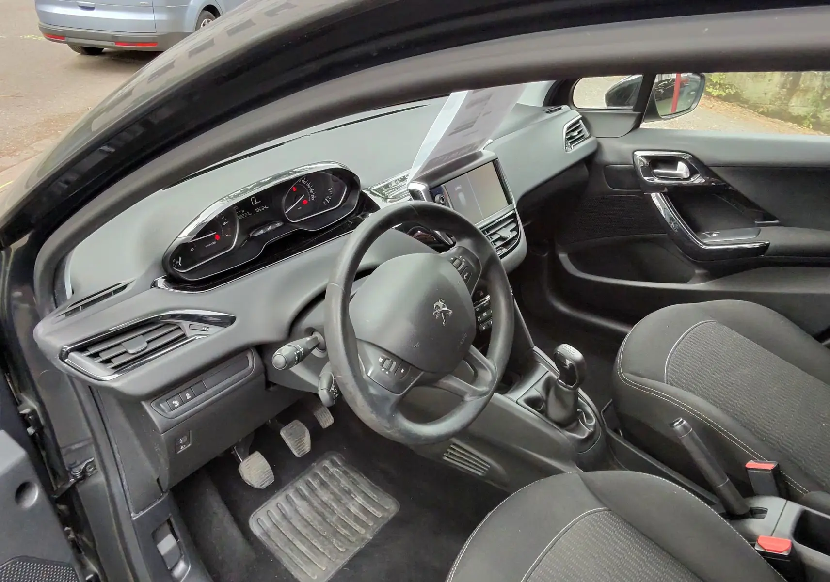 Vue intérieure côté conducteur de la Peugeot 208 gris foncé, montrant volant compact, tableau de bord numérique et sièges tissu noir.