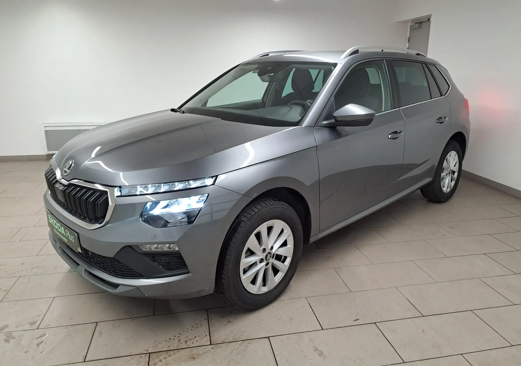 Vue 3/4 avant droite du Skoda Kamiq gris graphite 2025 avec phares allumés et caméra de recul intégrée.