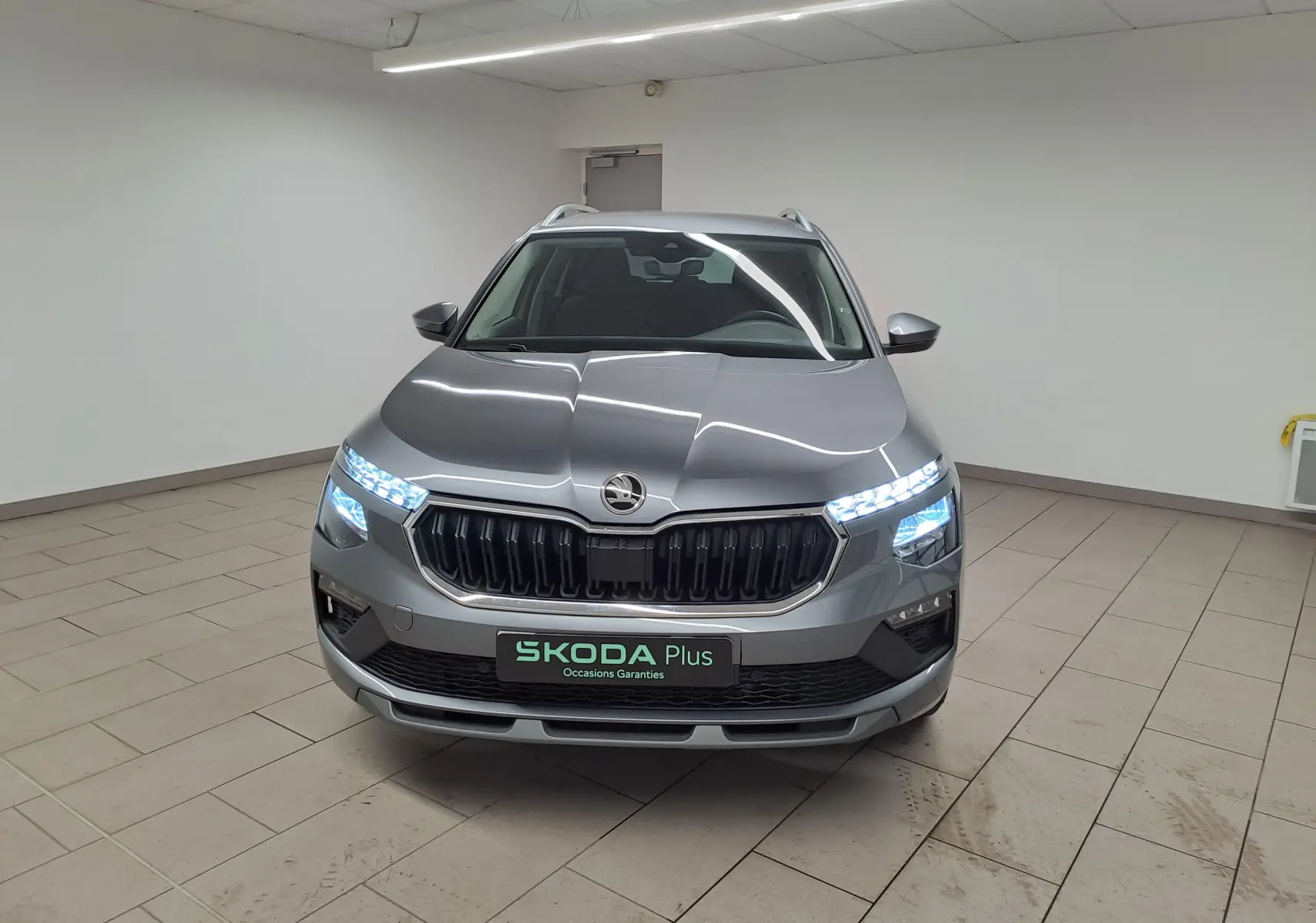 Vue avant d'un Skoda Kamiq gris graphite 2025 avec phares LED allumés dans un showroom intérieur.