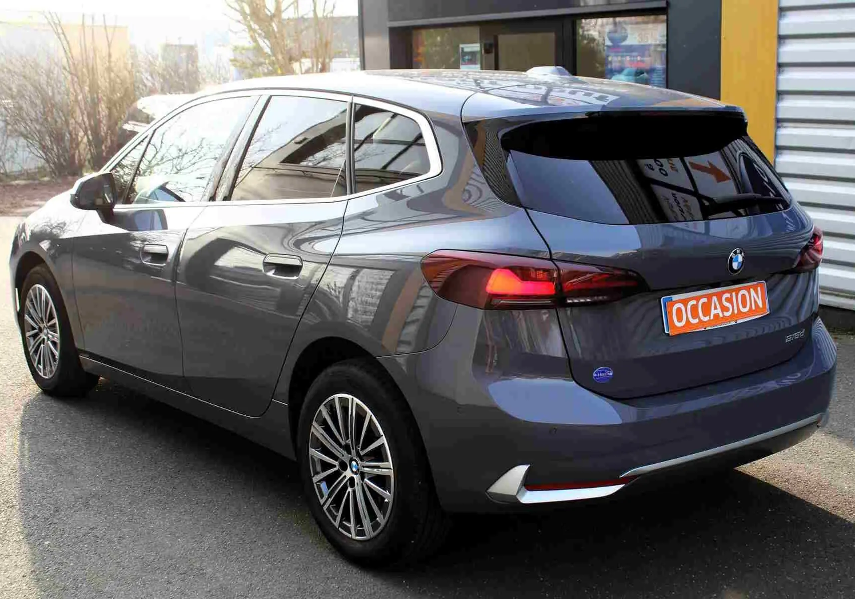 BMW Série 2 Active Tourer gris Nardo métal vue 3/4 arrière droit, feux arrière allumés et jantes alliage visibles.