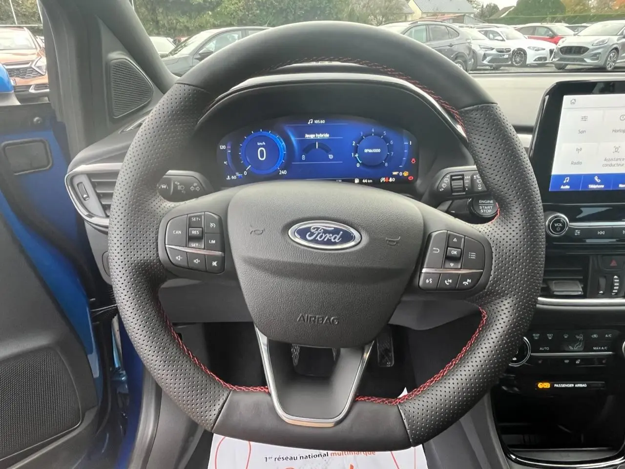 Vue intérieure centrée sur le volant perforé noir avec surpiqûres rouges d’un Ford Puma bleu, tableau de bord numérique visible.