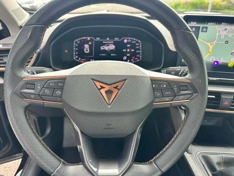Volant sport en cuir noir avec logo CUPRA cuivré, tableau de bord numérique et écran tactile GPS en arrière-plan.