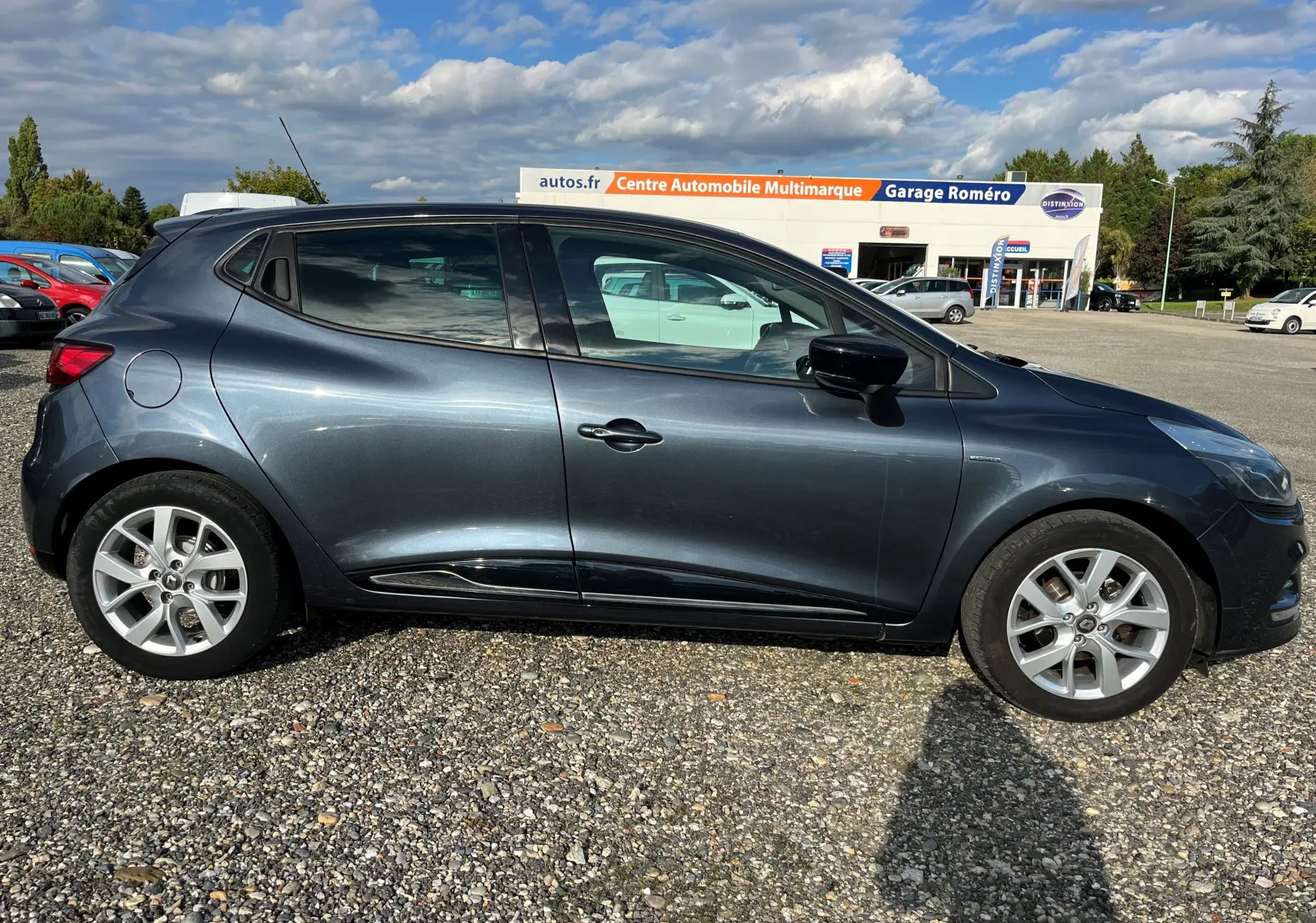 Profil côté gauche d'une Renault Clio IV gris foncé 2019 garée sur un sol gravillonné devant un garage multimarque.