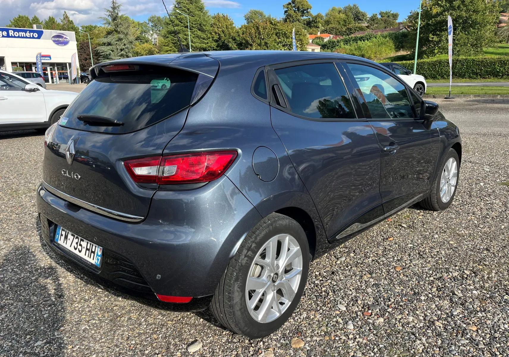Vue 3/4 arrière droite d'une Renault Clio IV gris métallisé garée sur un sol gravillonné par temps ensoleillé.