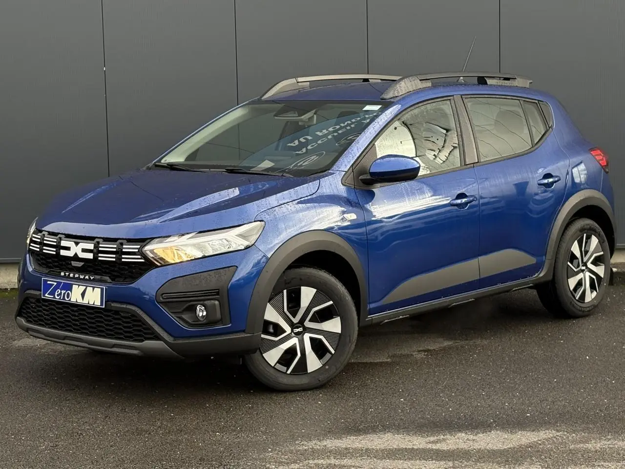 Vue 3/4 avant droite d'une Dacia Sandero Stepway 2025 Bleu Iron avec barres de toit et jantes spécifiques.