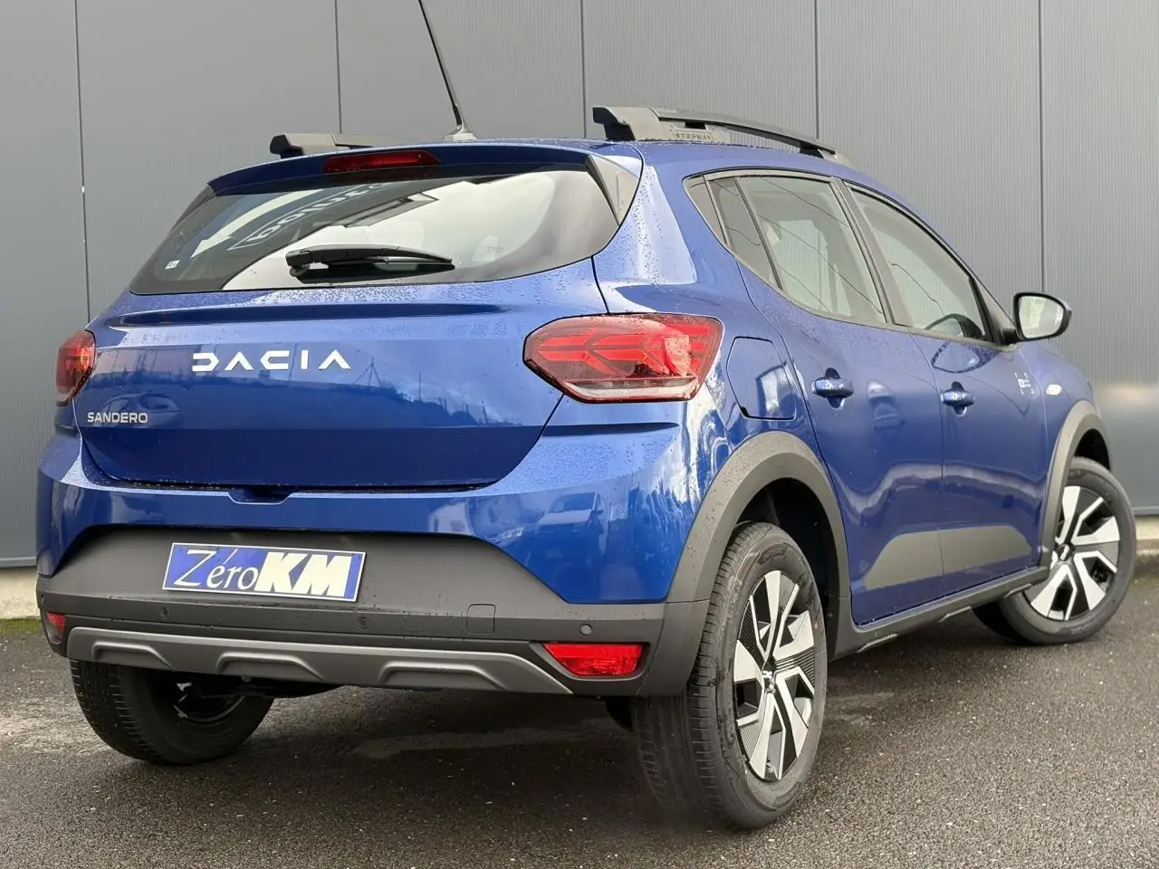 Vue 3/4 arrière droite d'une Dacia Sandero Stepway 2025 bleu Iron avec barres de toit et feux arrière LED en Y.