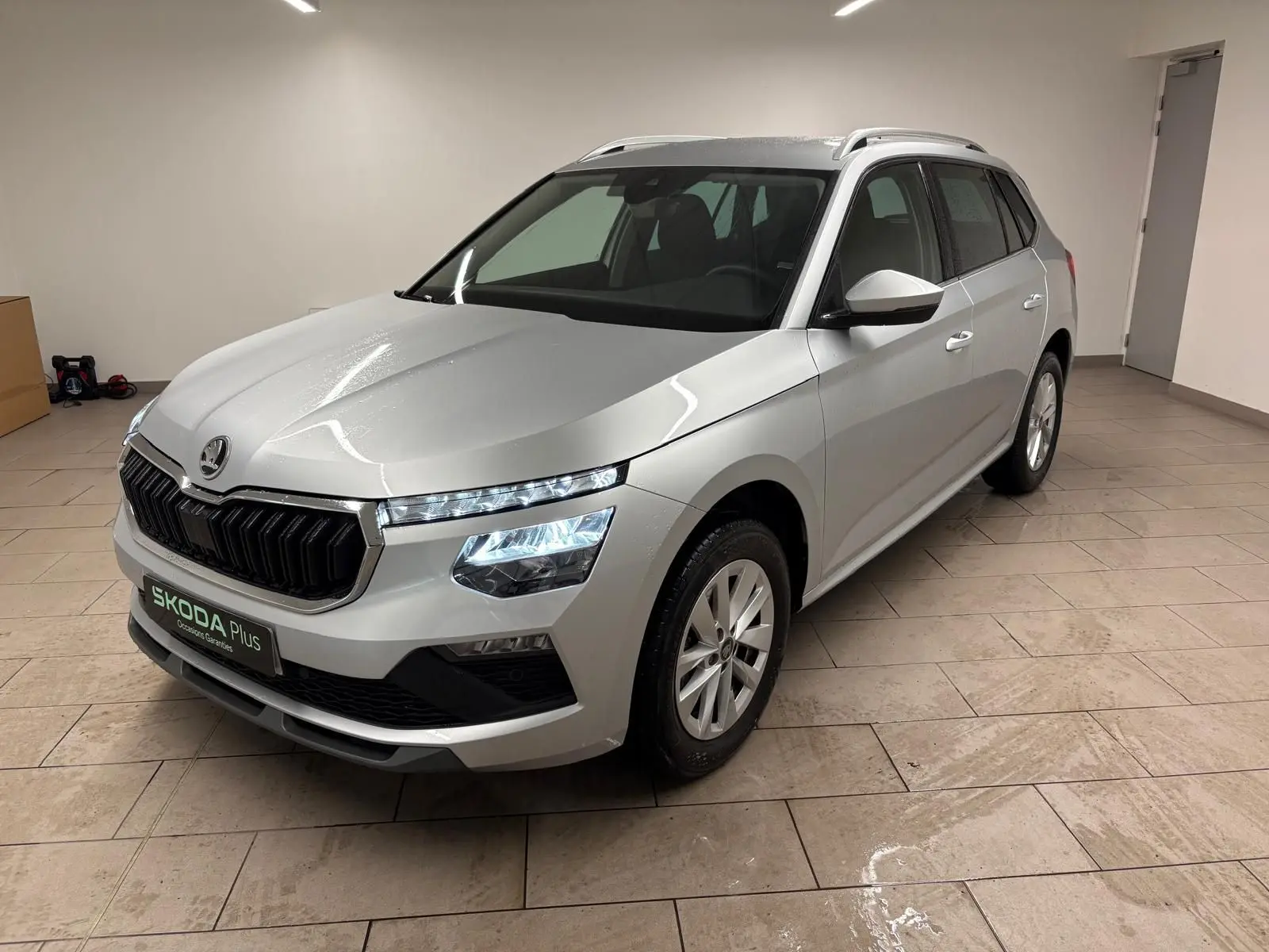 Vue 3/4 avant d'un Skoda Kamiq gris argent métallisé, avec phares allumés et calandre noire distinctive.