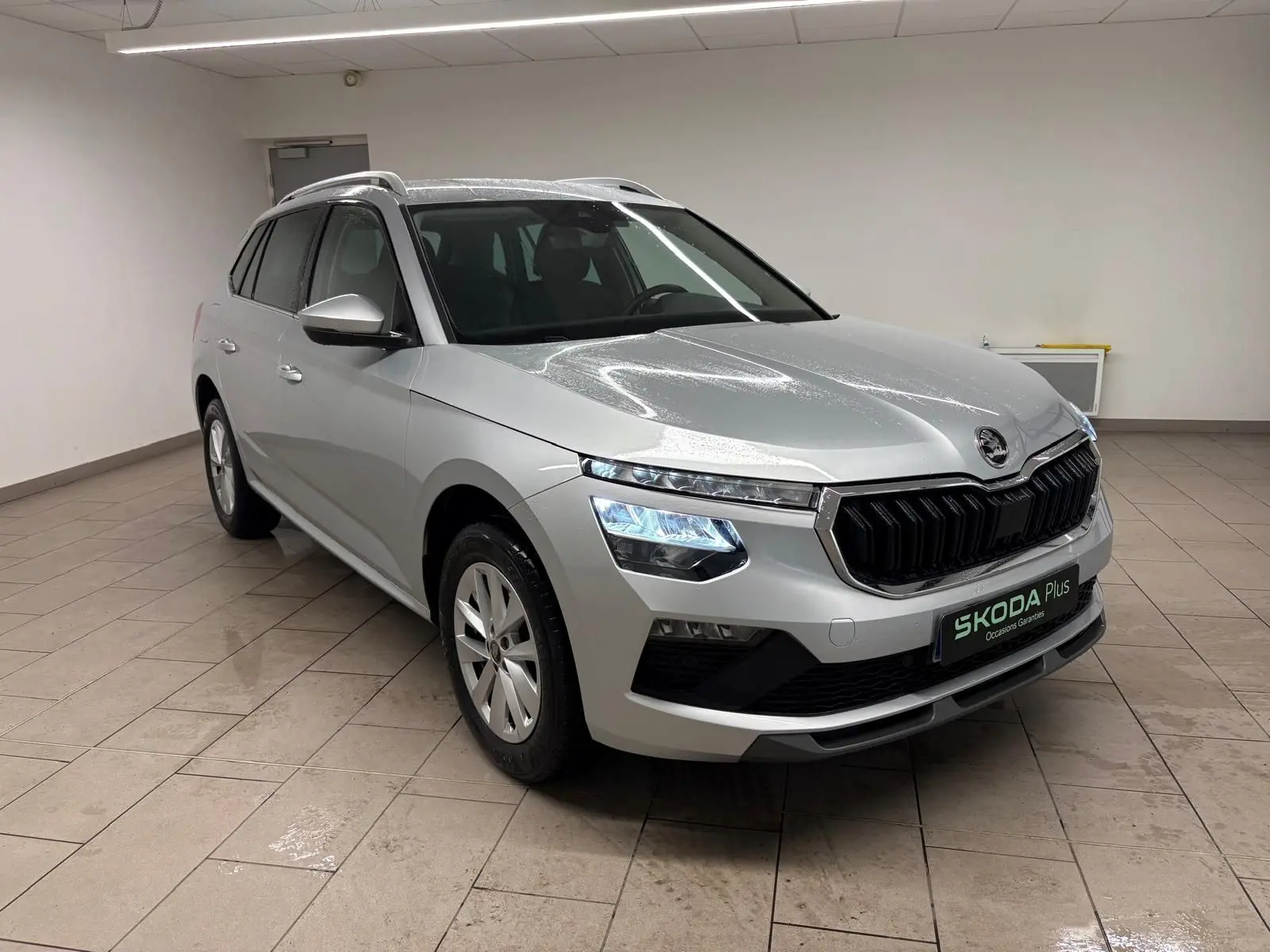 Vue 3/4 avant d'un Skoda Kamiq gris argent métallisé avec calandre noire et phares LED allumés, en intérieur.