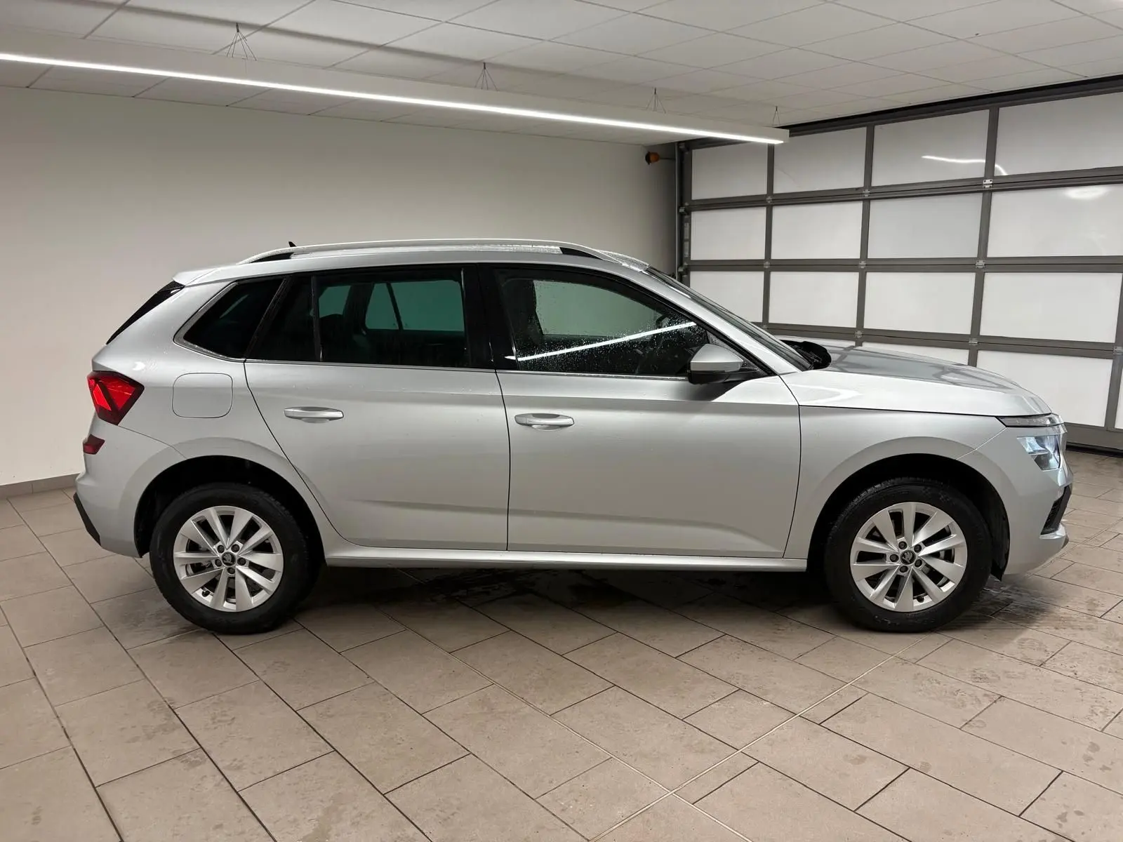 Vue de profil côté gauche d'un Skoda Kamiq gris argent métallisé dans un garage, avec caméra de recul visible.