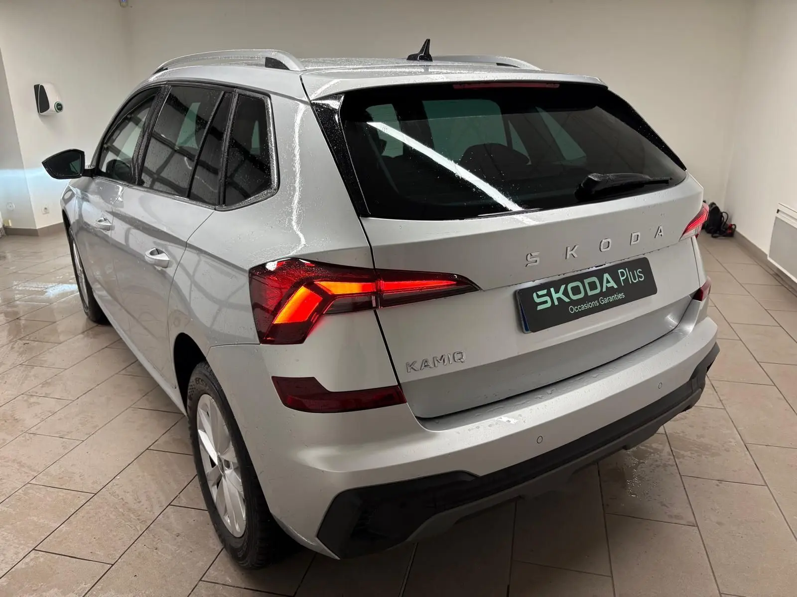 Vue 3/4 arrière gauche du Skoda Kamiq gris argent 2025 avec feux arrière allumés et caméra de recul visible.