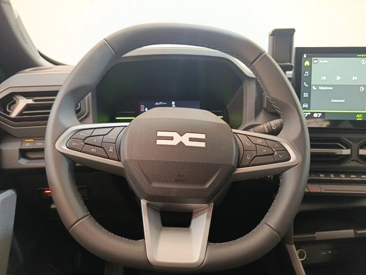 Vue rapprochée du volant noir multifonctions du Dacia Duster Journey Hybrid 155 avec tableau de bord et écran tactile visible.