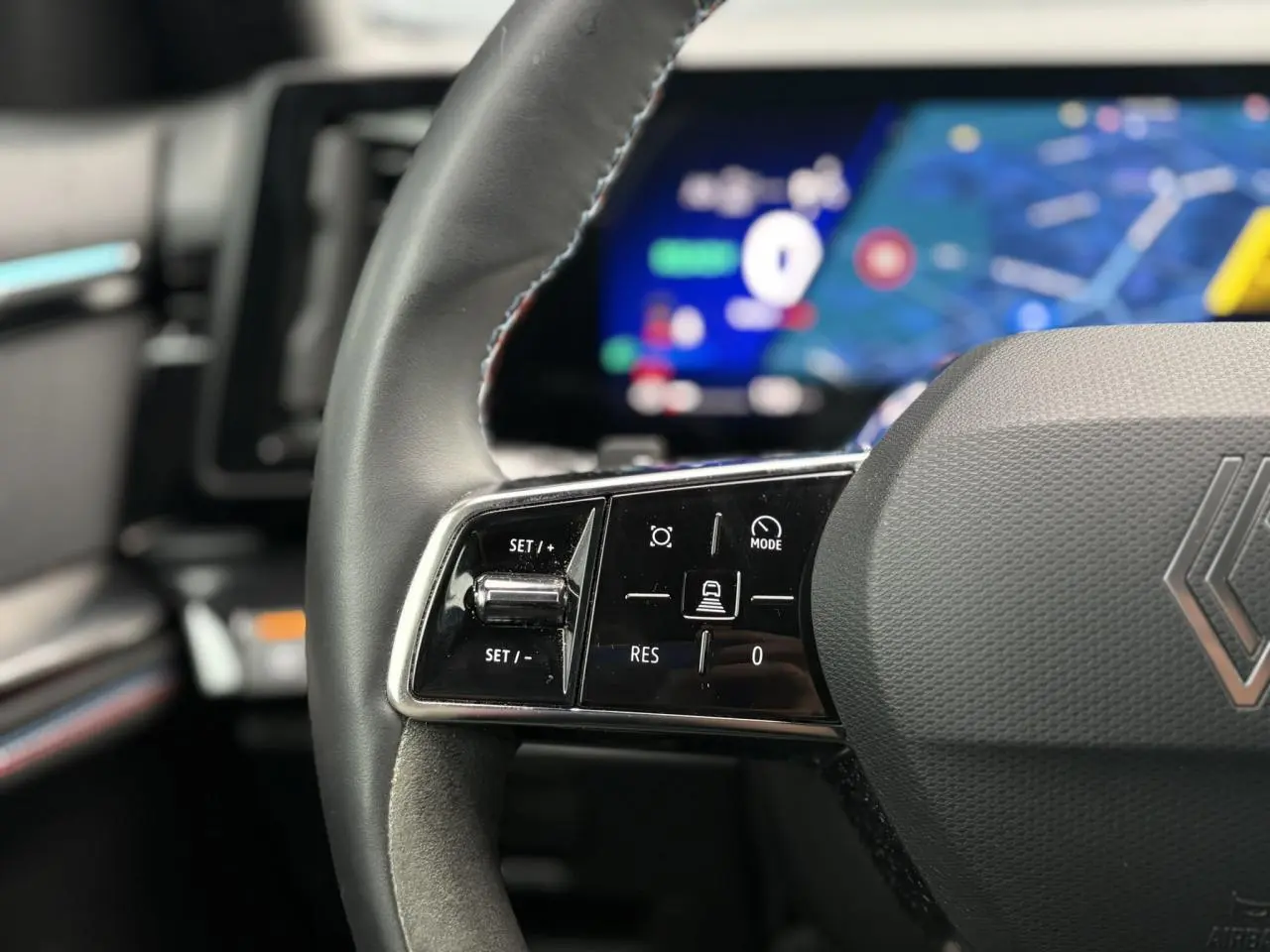 Gros plan sur le volant à droite du Renault Rafale Bleu Sommet, avec commandes intégrées et tableau de bord numérique flou en arrière-plan.