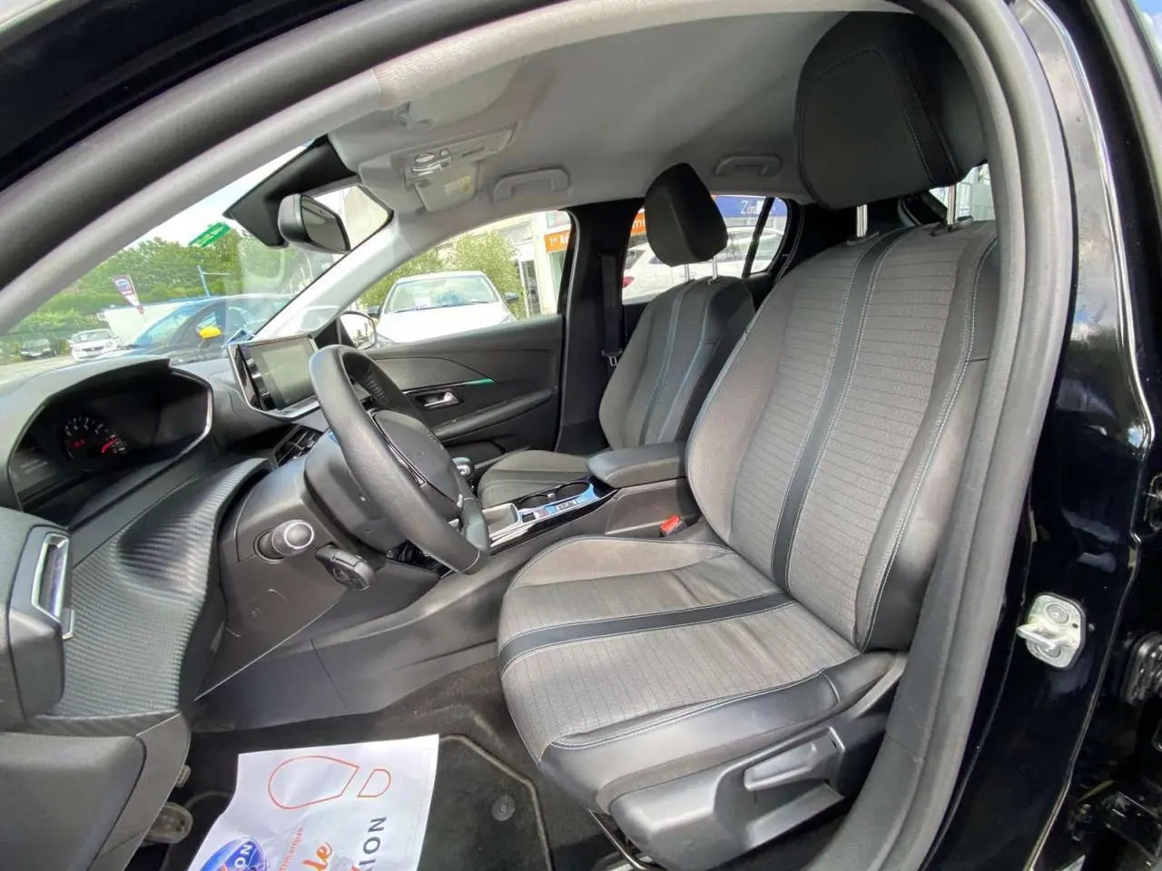 Vue intérieure côté conducteur de la Peugeot 208 noire, mettant en valeur les sièges gris clair et le tableau de bord moderne.
