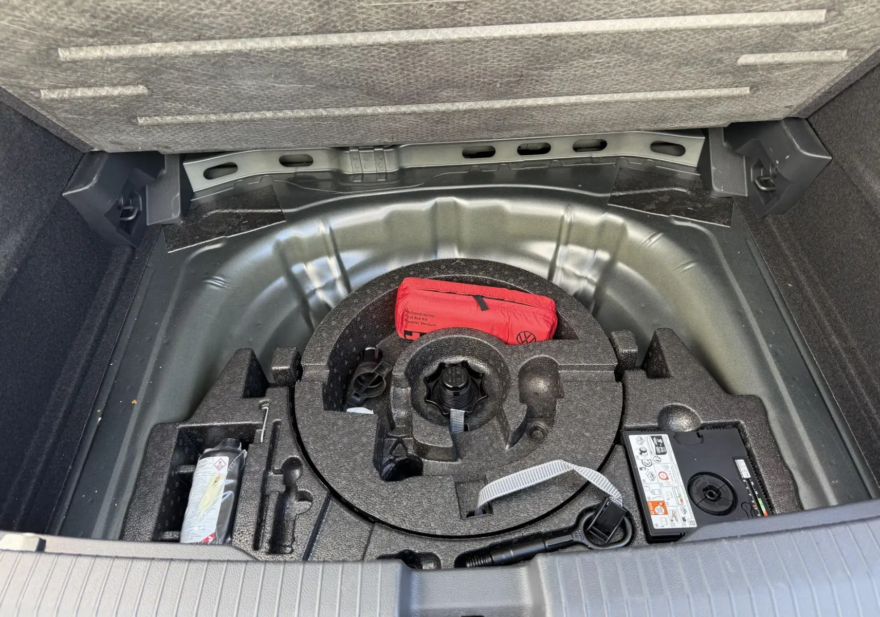 Vue du coffre ouvert d'un Volkswagen T-Roc gris, montrant le rangement sous plancher avec kit de réparation et trousse de secours rouge.