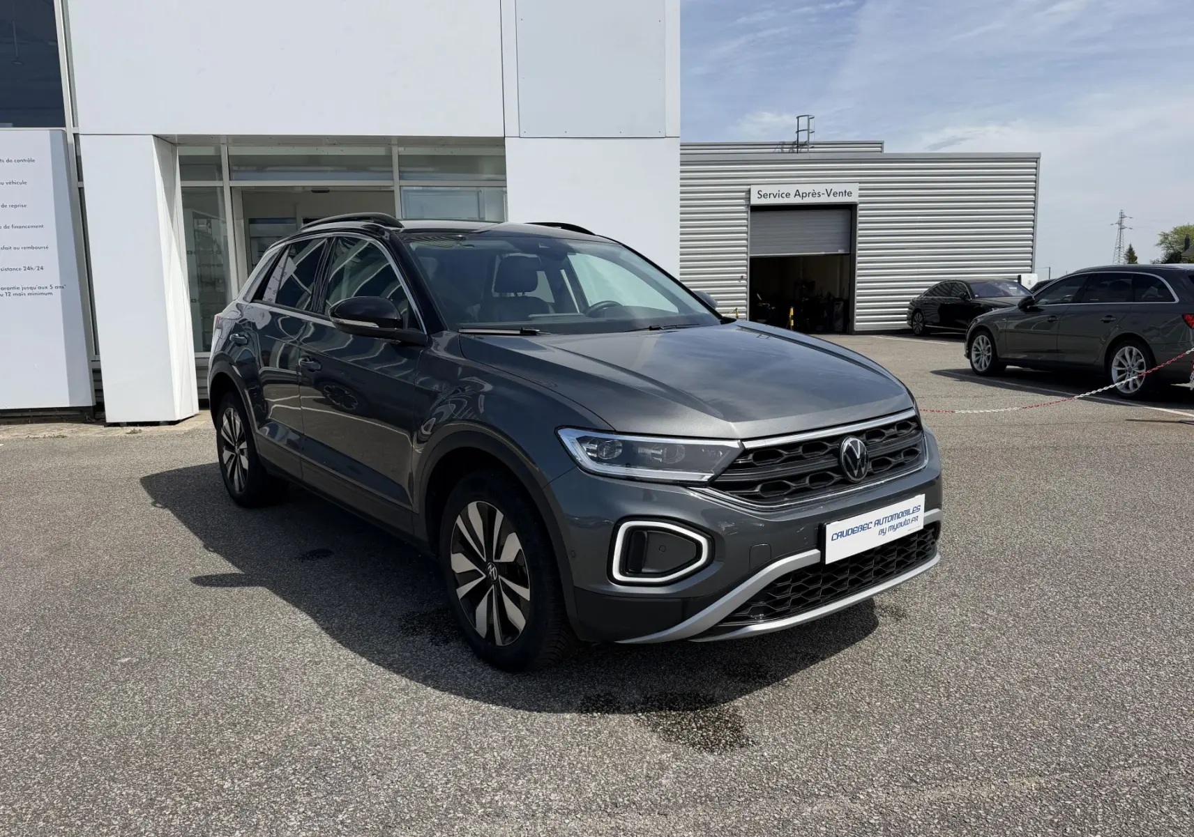 Volkswagen T-Roc gris en 3/4 avant droit, avec barres de toit noires et jantes au design moderne, stationné devant un bâtiment.