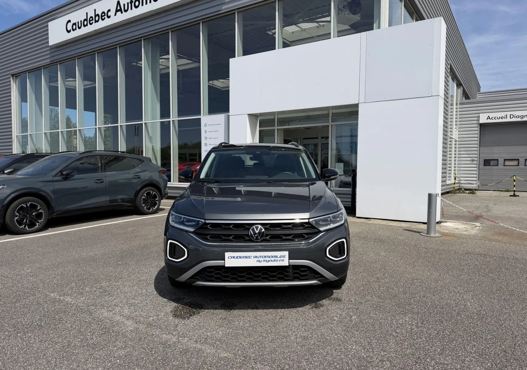 Vue de face du Volkswagen T-Roc gris 2025 avec calandre noire et feux LED en forme de C, devant un showroom.