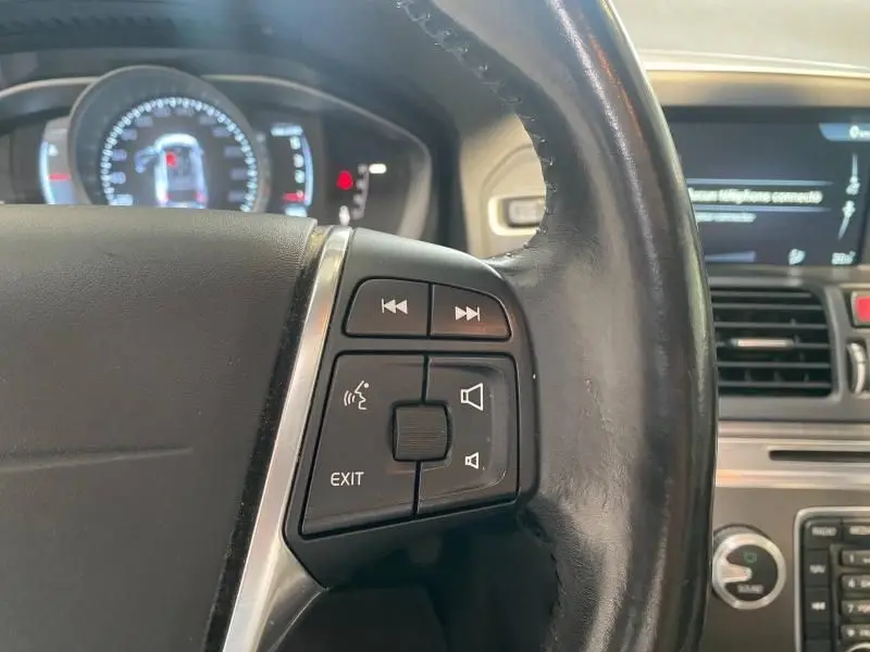 Gros plan sur les commandes audio et vocales au volant cuir du Volvo XC60 argenté, tableau de bord en arrière-plan flou.
