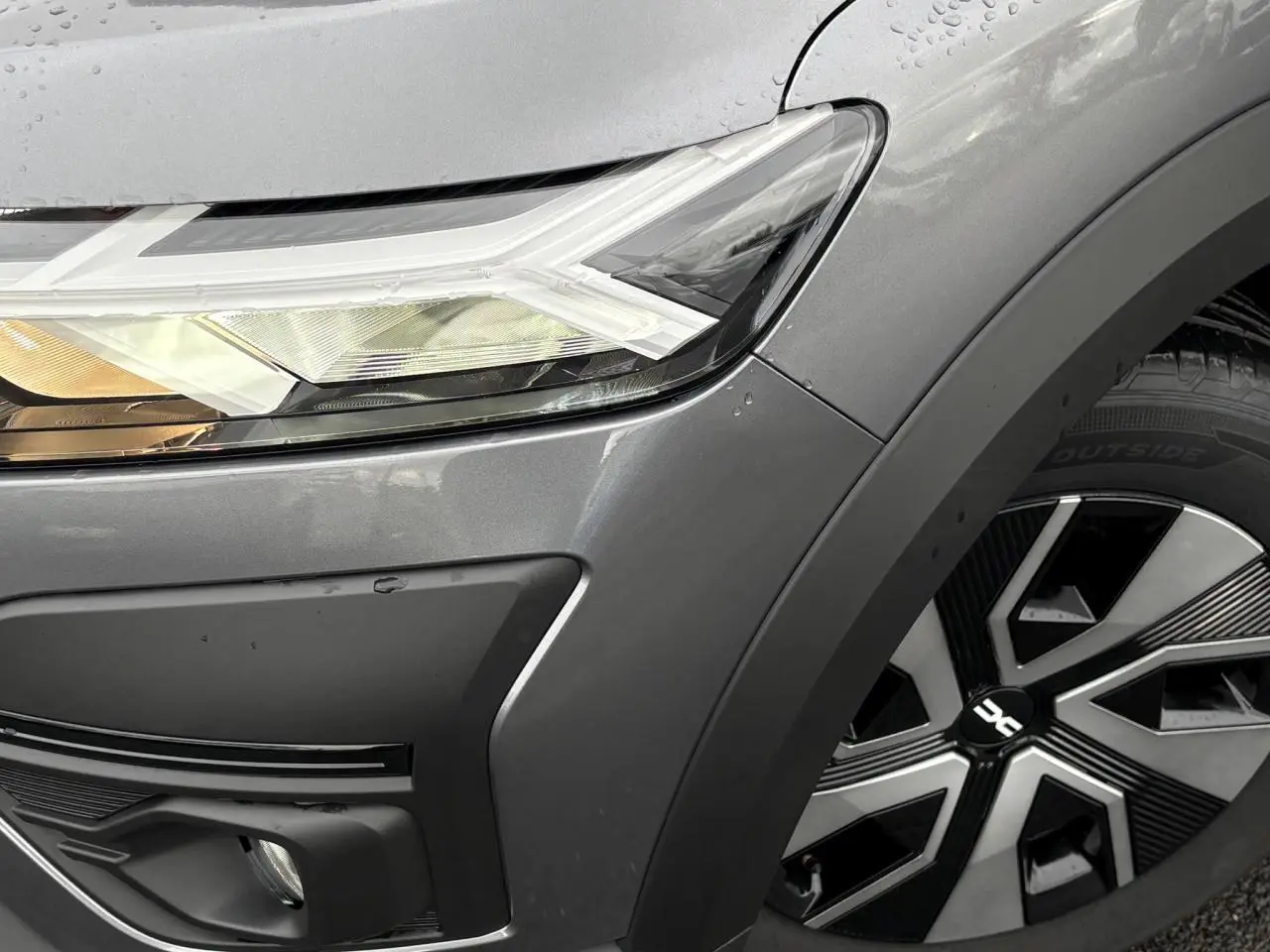 Gros plan sur le phare avant gauche et la roue avec enjoliveur noir et argent d'une Dacia Sandero Stepway gris schiste.