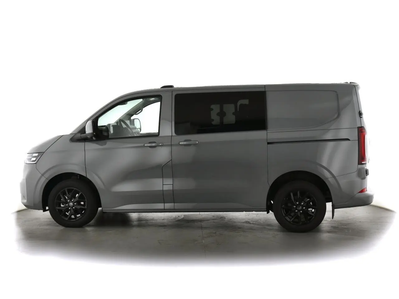 Vue de profil côté gauche d’un Volkswagen Transporter T7 gris Stone avec jantes alliage noir brillant et vitres arrière surteintées.