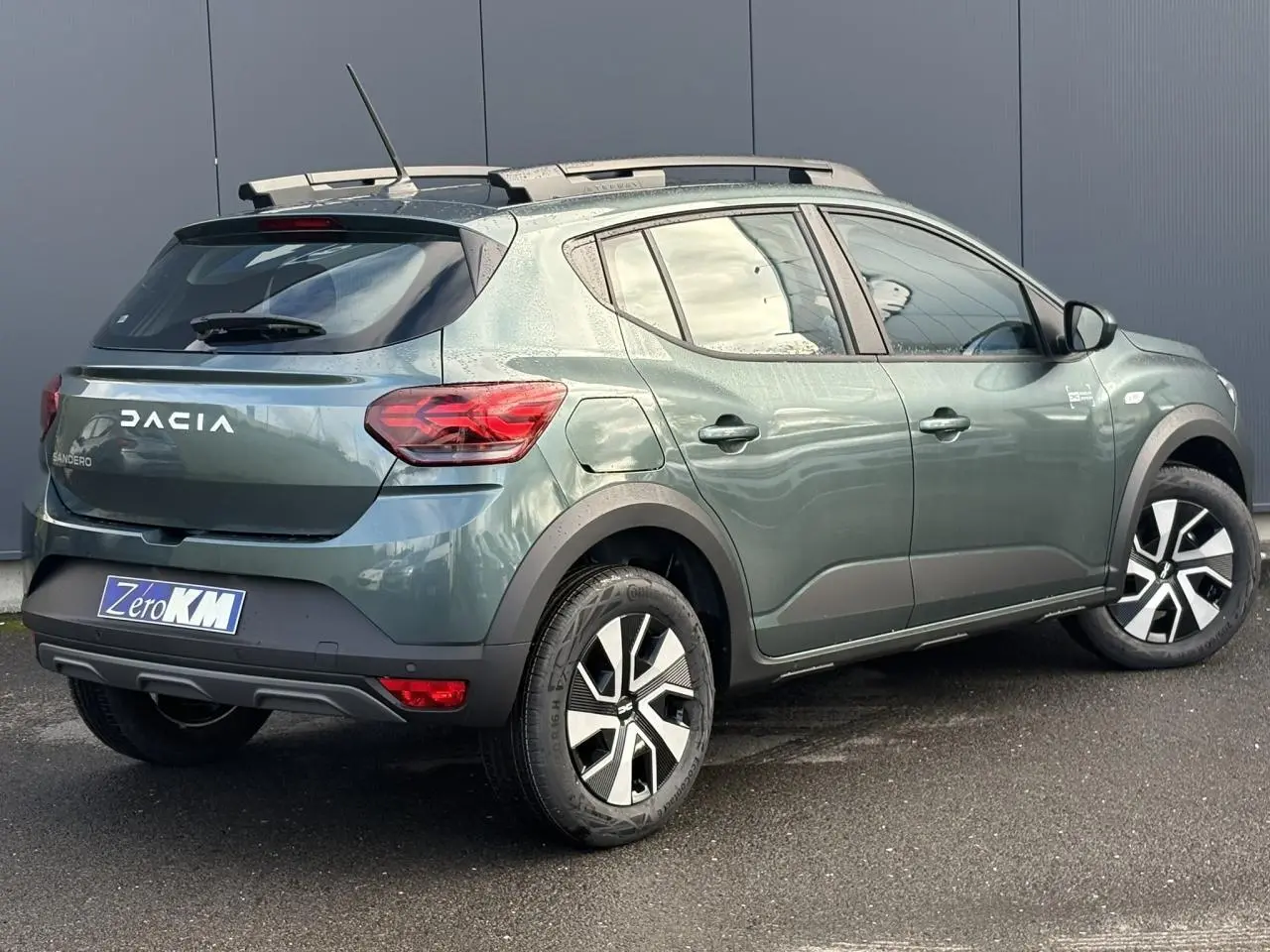 Vue 3/4 arrière droite d'une Dacia Sandero Stepway 2025 vert cèdre avec barres de toit et jantes spécifiques.