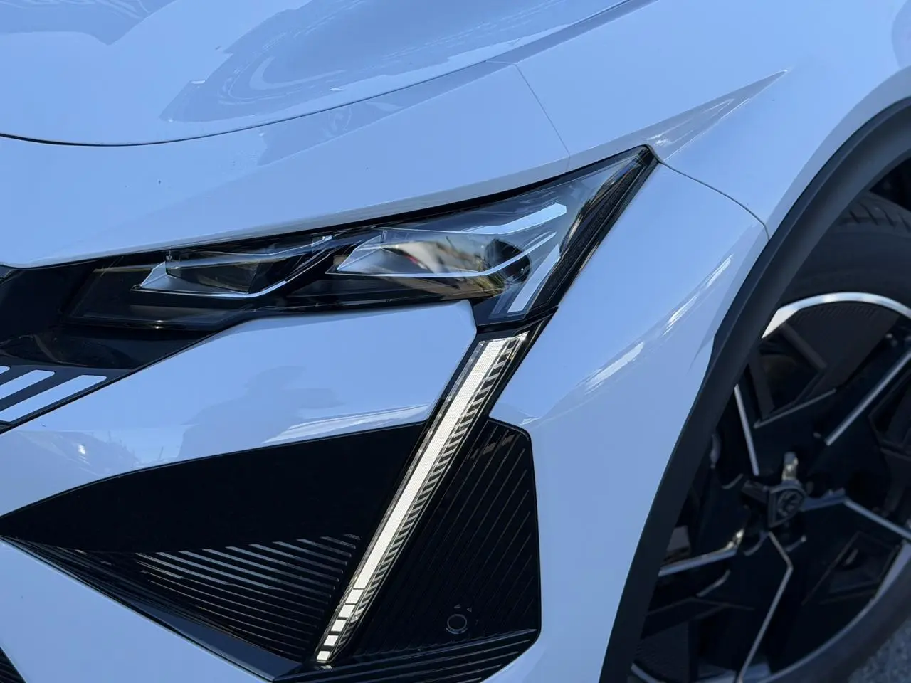 Gros plan sur le phare avant droit blanc Okenite du Peugeot 408 Hybrid 145 e-DSC6 GT avec jante alliage noire diamantée.