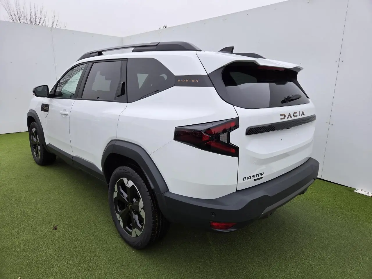 Vue 3/4 arrière droite du Dacia Bigster blanc glacier avec jantes alliage et détails brun cuivré sur fond vert.