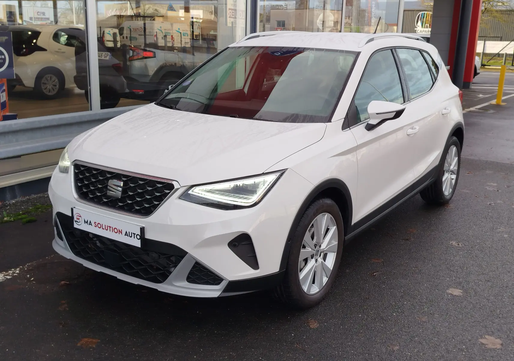 SEAT Arona blanc candy vue 3/4 avant droit, avec calandre noire et feux LED allumés, stationné devant un bâtiment vitré.