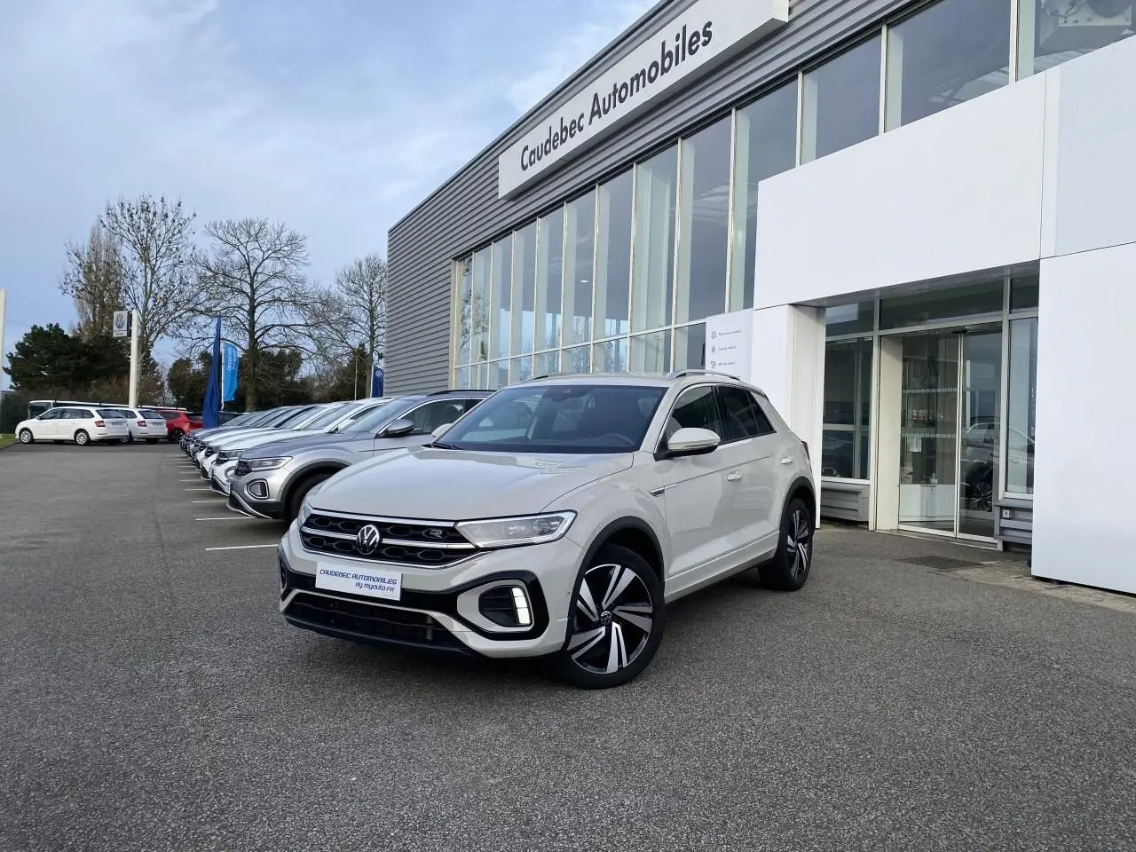 Volkswagen T-Roc gris en 3/4 avant droit, avec jantes alliage noires et calandre R-Line visible devant un concessionnaire.