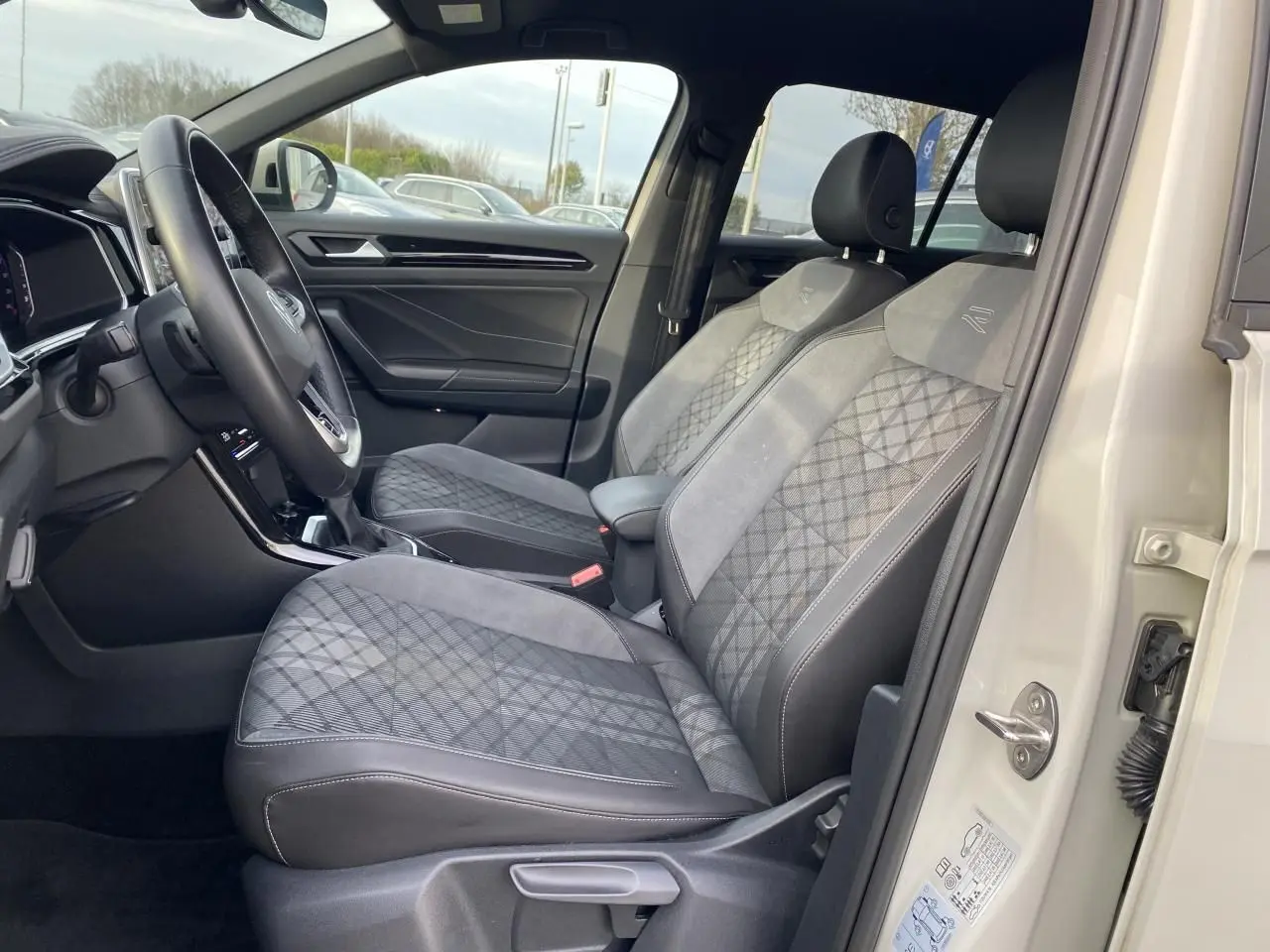 Vue intérieure côté conducteur du Volkswagen T-Roc 2024, sièges gris à motifs et volant multifonction en cuir.