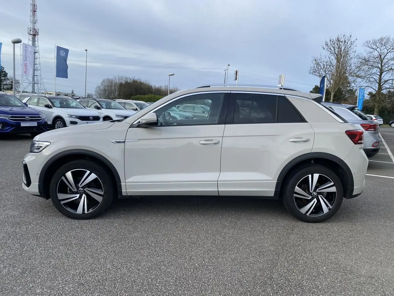 Vue de profil côté gauche d'un Volkswagen T-Roc gris clair avec jantes alliage bicolores sur un parking extérieur.