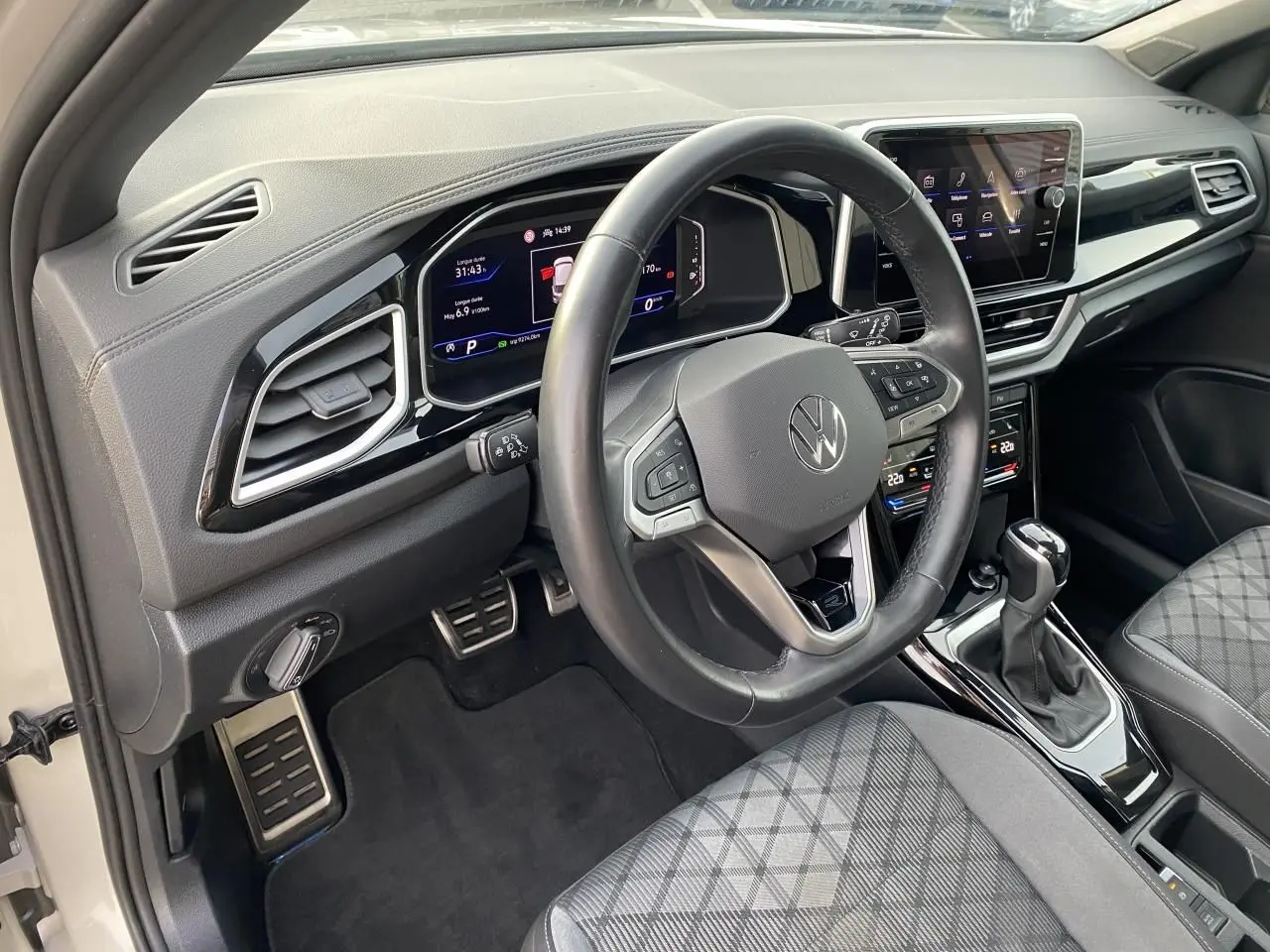 Vue intérieure côté conducteur du Volkswagen T-Roc 2024, volant cuir noir, tableau de bord digital et sièges tissu gris à motif.