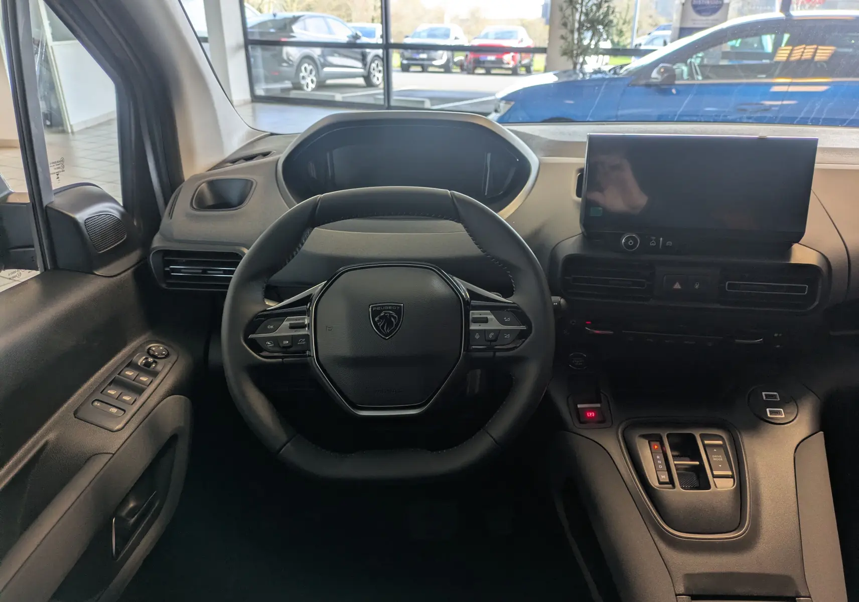 Vue intérieure centrée sur le volant noir du Peugeot Rifter 2025, avec tableau de bord et écran tactile en arrière-plan.