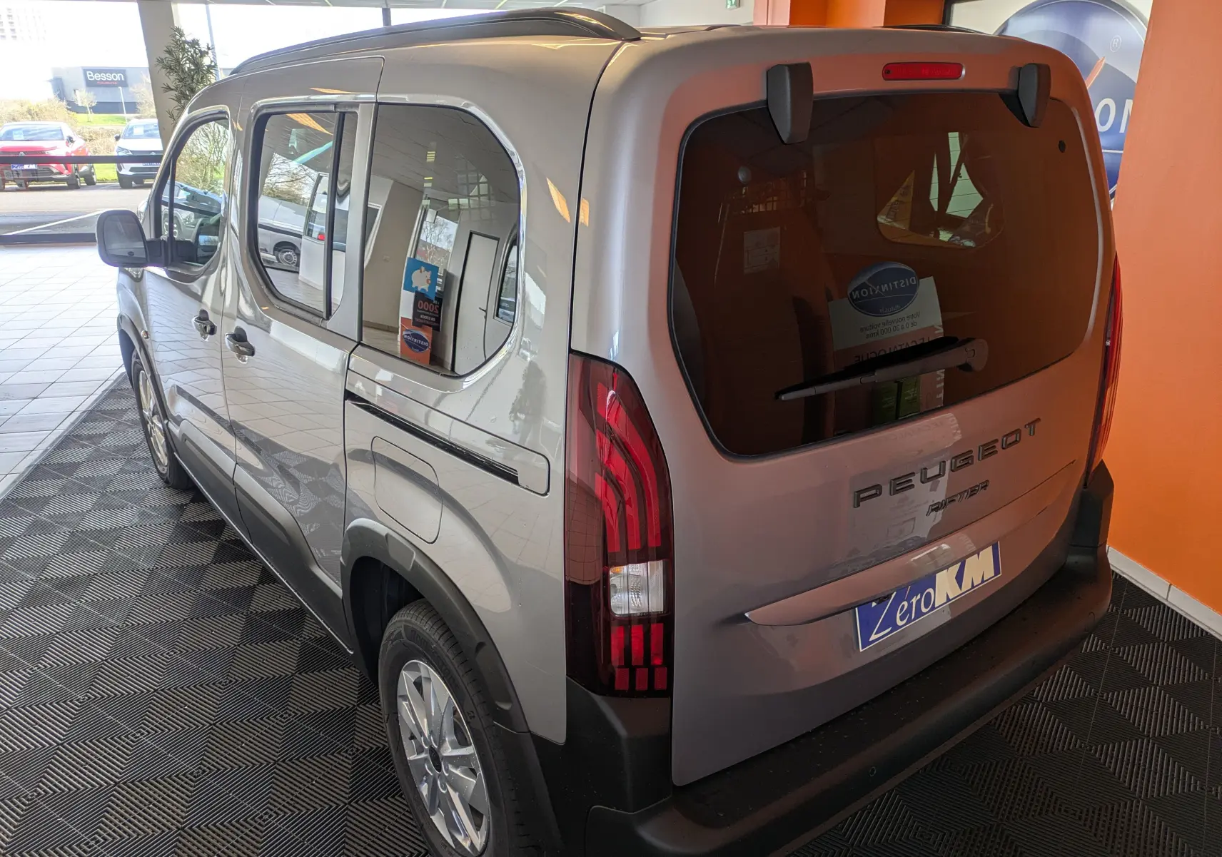 Vue 3/4 arrière gauche du Peugeot Rifter gris Artense 2025 avec barres de toit noir brillant et vitres teintées.