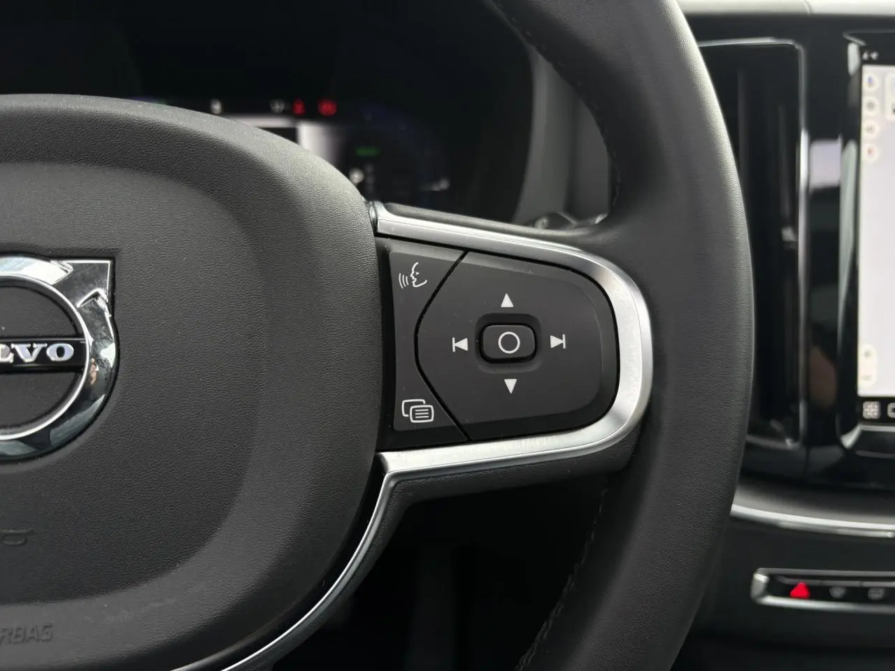 Gros plan sur le volant simili-cuir noir du Volvo XC60 2024, avec commandes multifonctions et écran tactile visible.