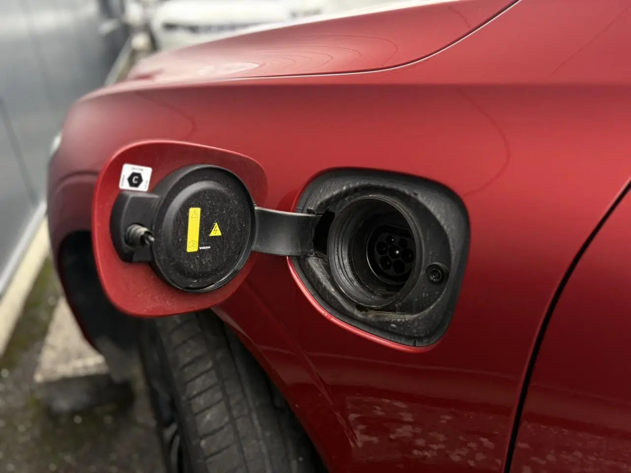 Prise de recharge ouverte côté gauche d’un Volvo XC60 T6 AWD PHEV 2024 rouge Fusion, détail du port de charge hybride.