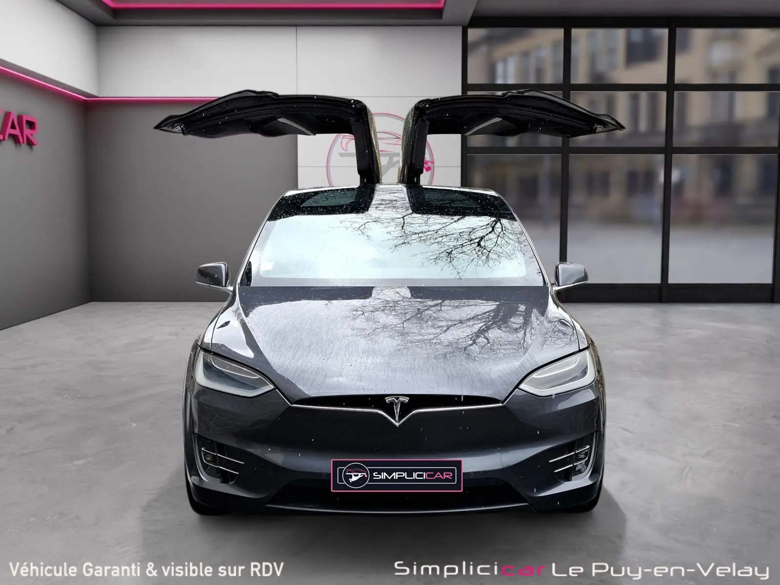 Vue frontale d’une Tesla Model X 100D gris foncé avec portes papillon ouvertes dans un showroom moderne.