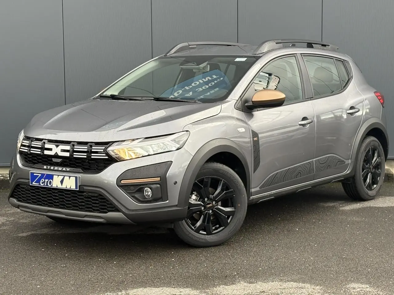 Dacia Sandero Stepway TCE 110 gris schiste en 3/4 avant droit avec jantes noires et barres de toit grises.