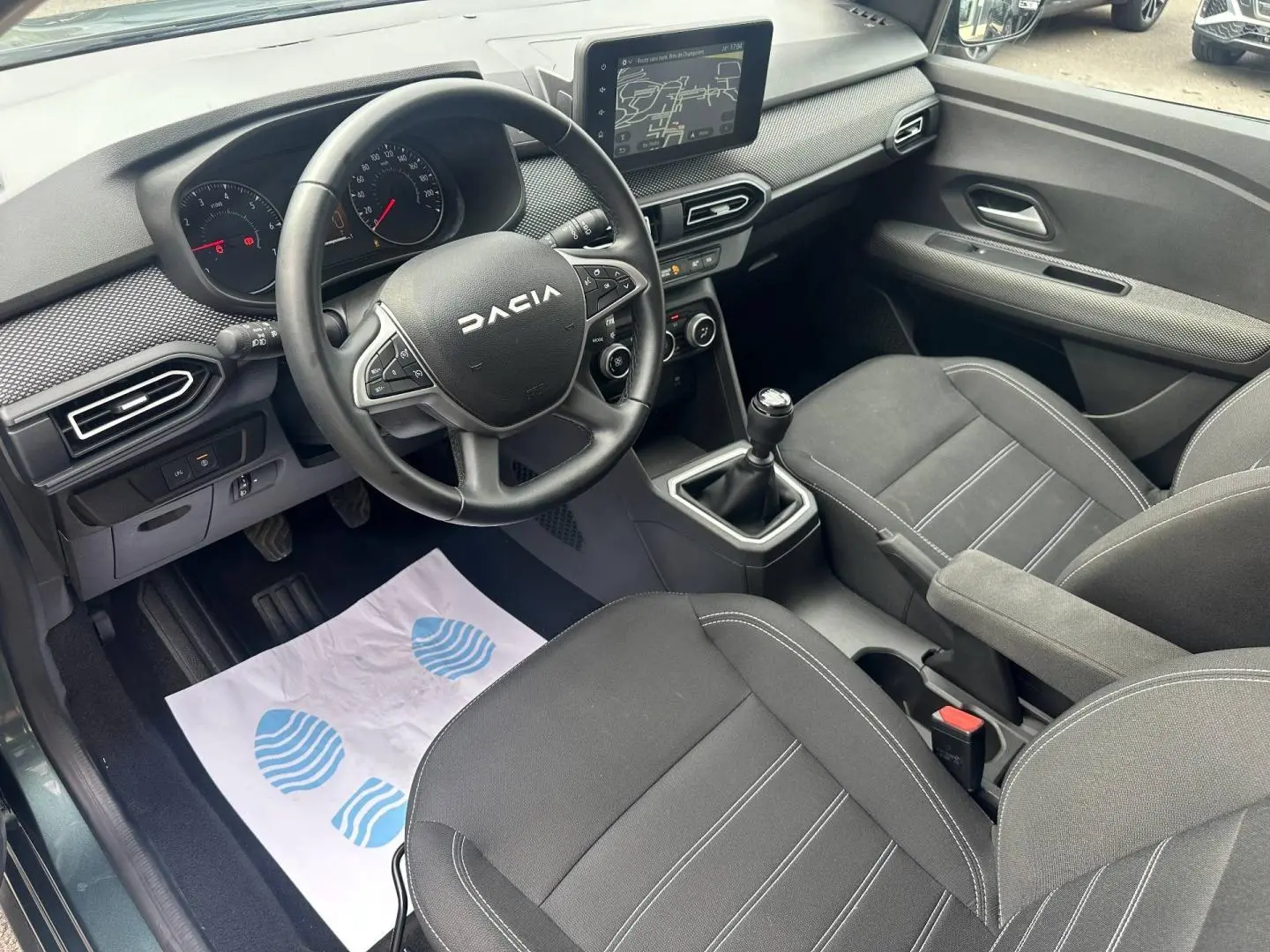 Vue intérieure côté conducteur du Dacia Jogger 2025, volant noir, tableau de bord avec écran tactile et sièges tissu gris foncé.