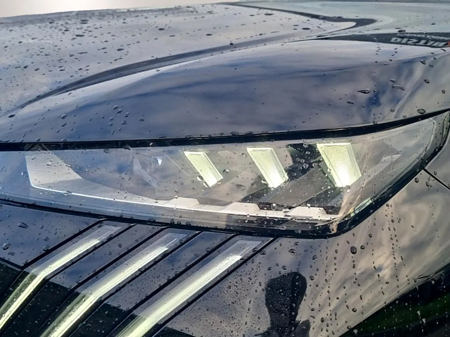 Gros plan sur le phare avant droit noir brillant du Peugeot 5008 Hybrid 145 GT avec pluie sur la carrosserie.